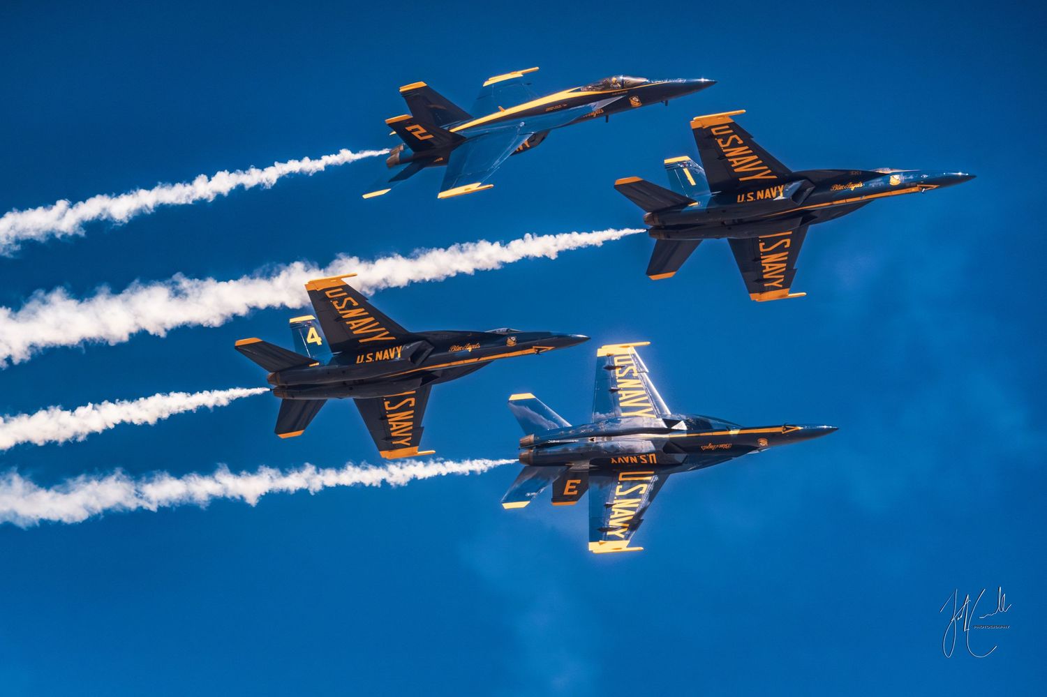 Four Blue Angels jets flying in formation with white smoke trails against bright blue sky.