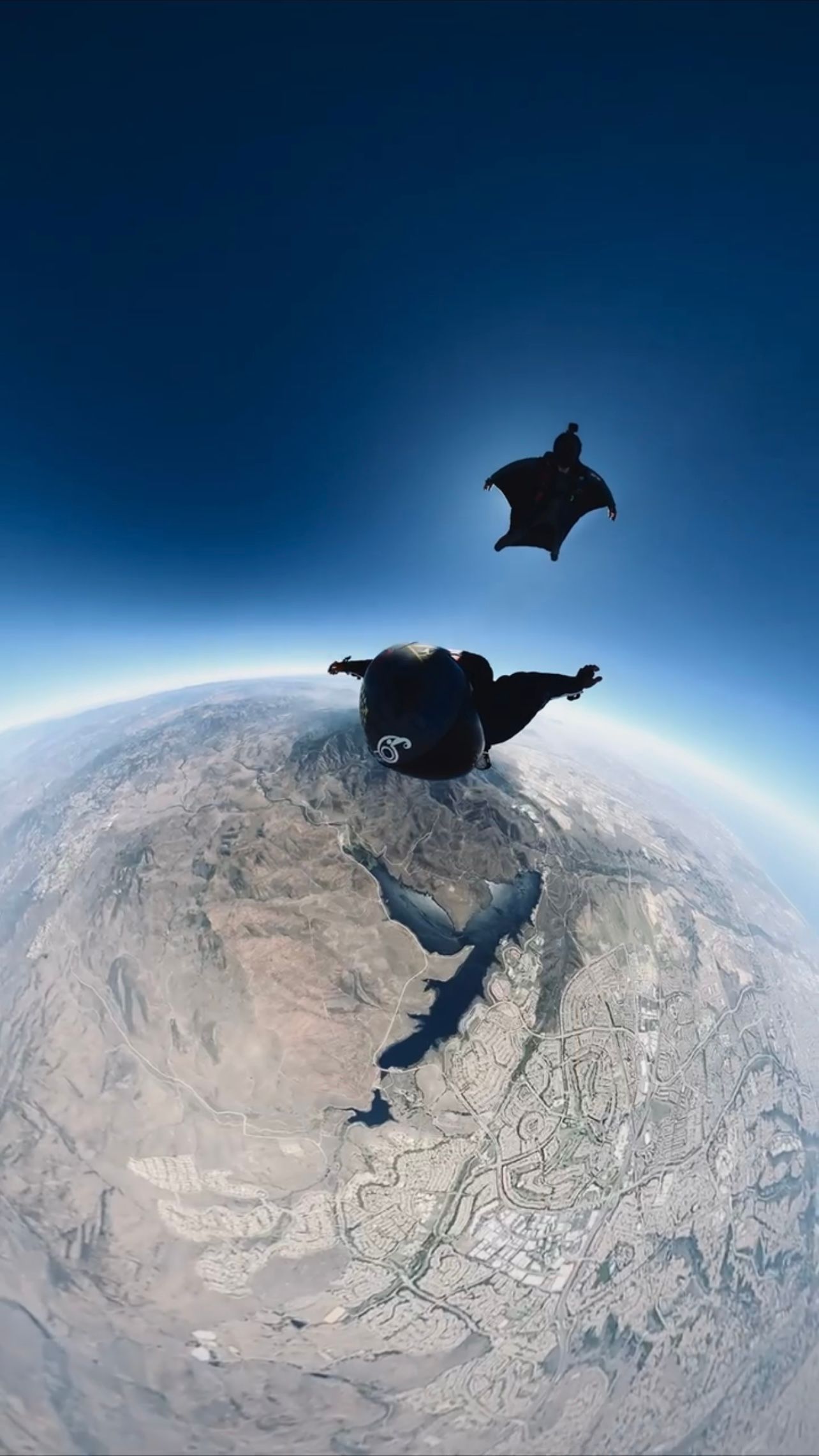 Two wingsuit flyers soar high above a mountainous landscape with curved horizon visible.