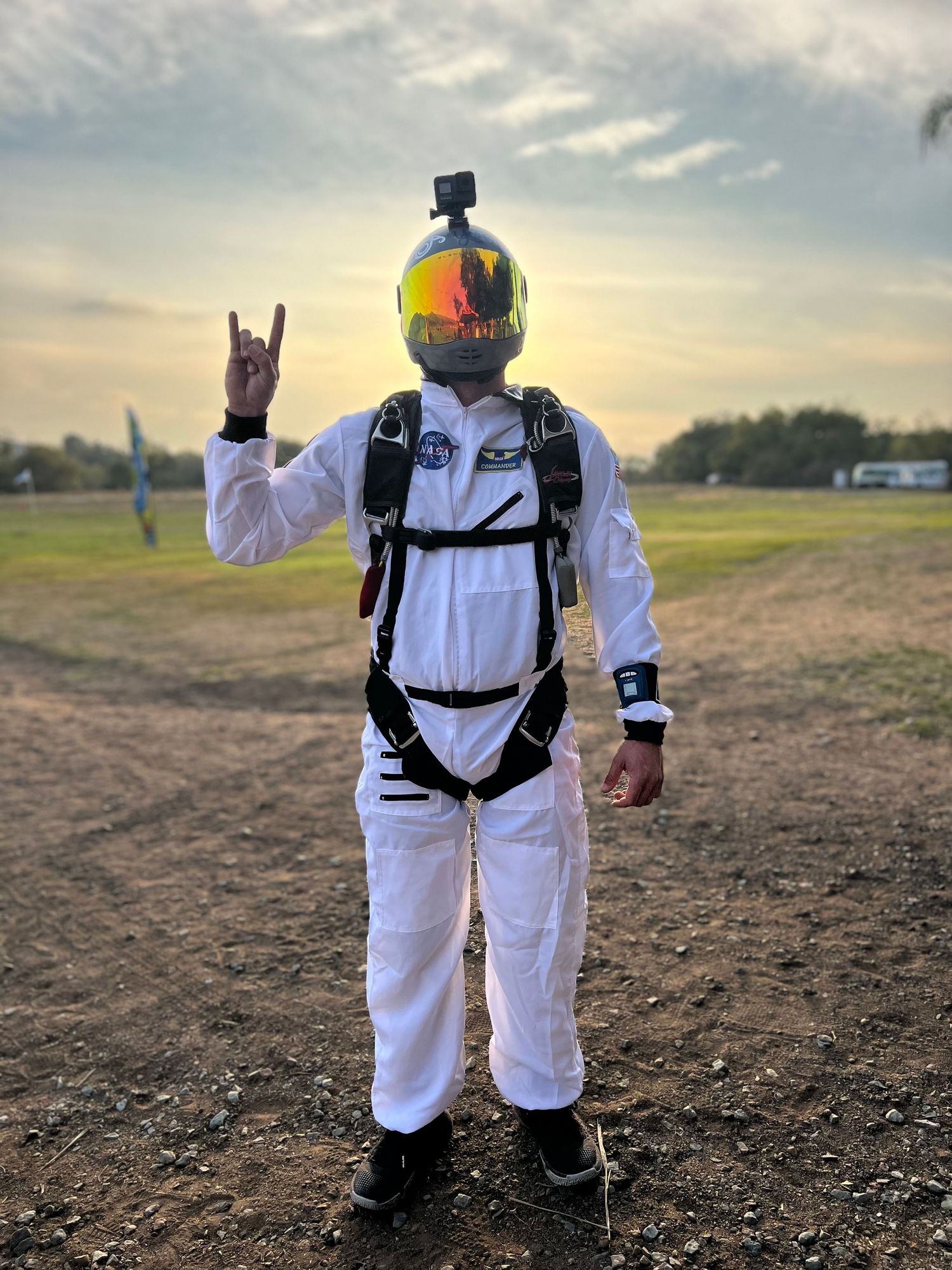 Skydiver in white jumpsuit and safety gear preparing for jump on a field at dusk.