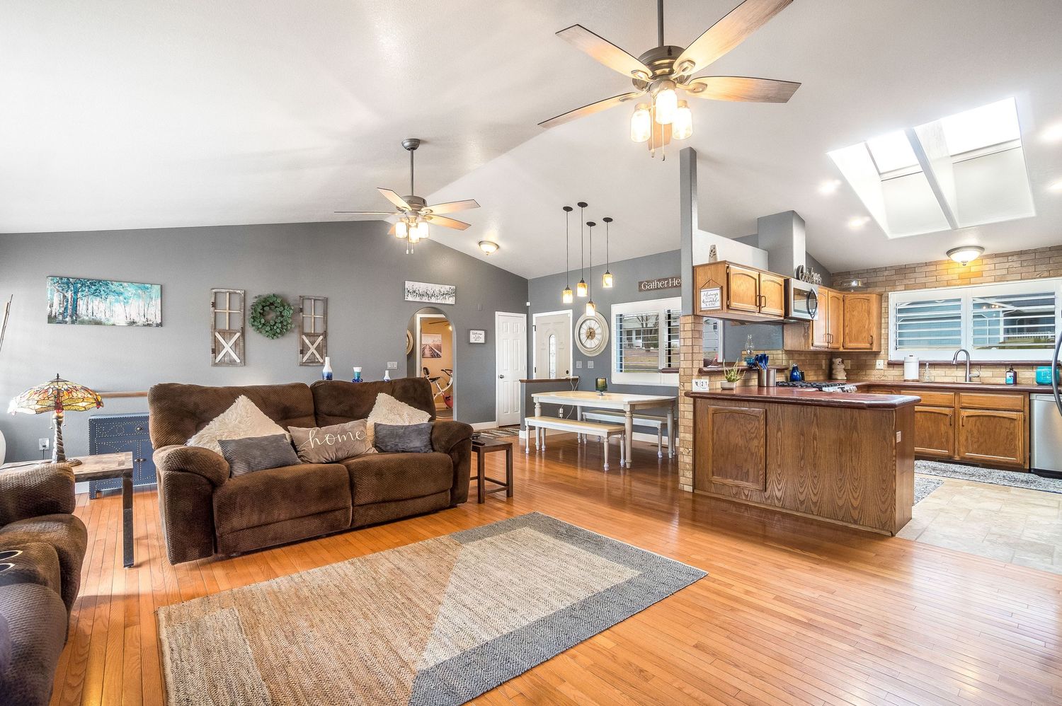 Open-concept living room with hardwood floors, vaulted ceiling, ceiling fan, and adjacent kitchen with wood cabinets.