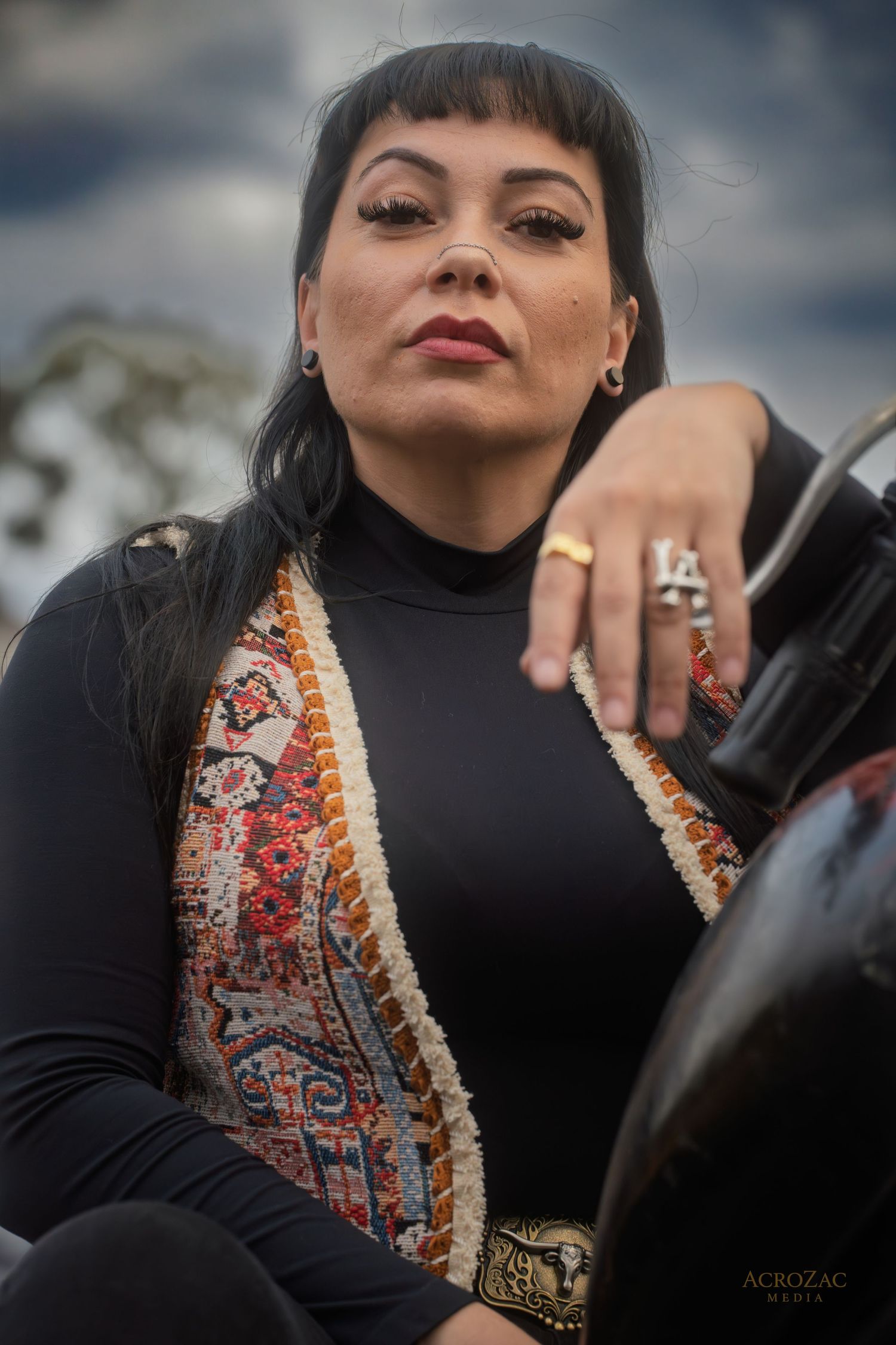 Portrait of someone wearing a black turtleneck and patterned vest with ornate rings against a blurred outdoor background.