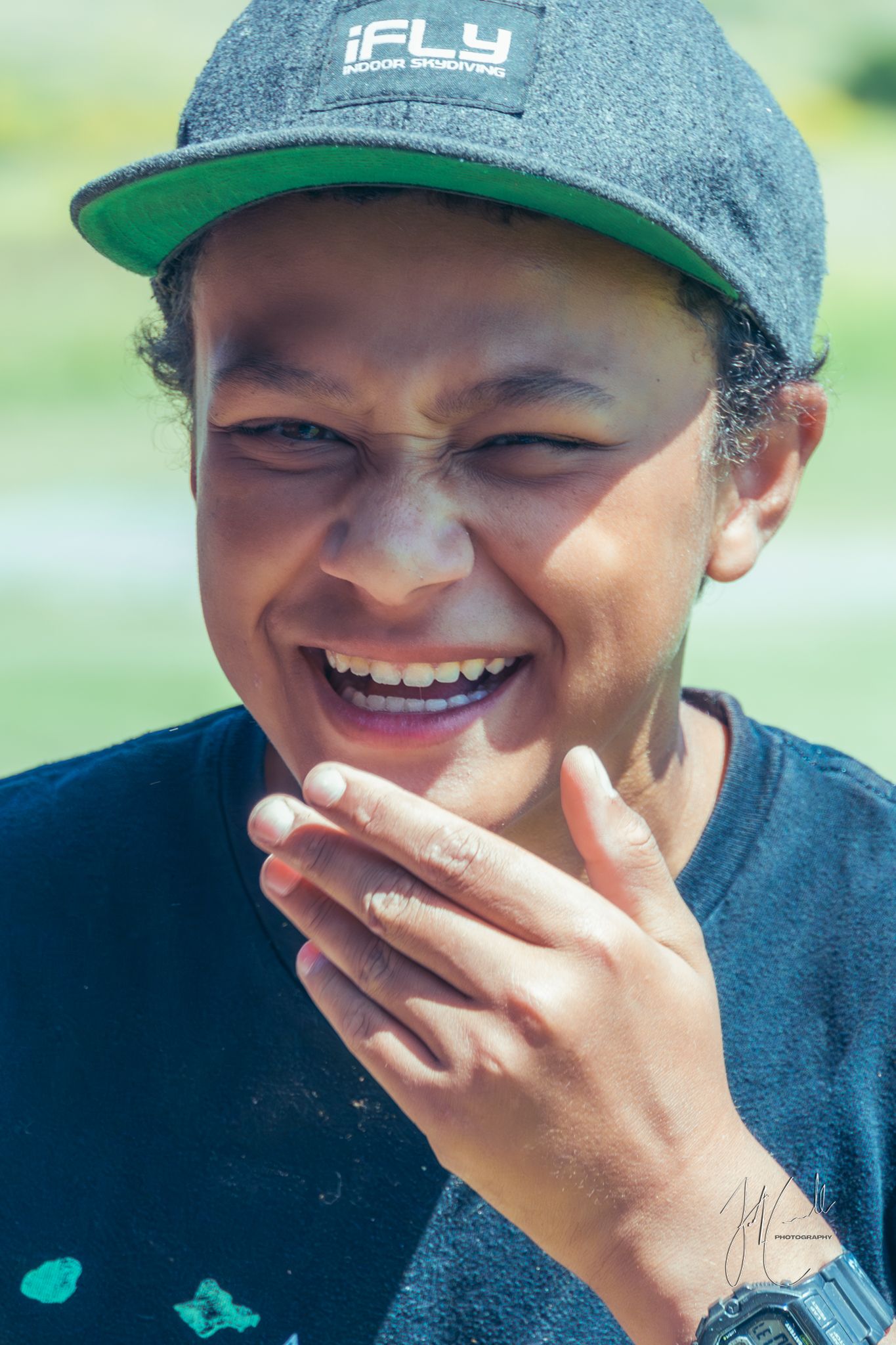 Person wearing a grey cap and iFLY shirt smiles warmly while covering their mouth.