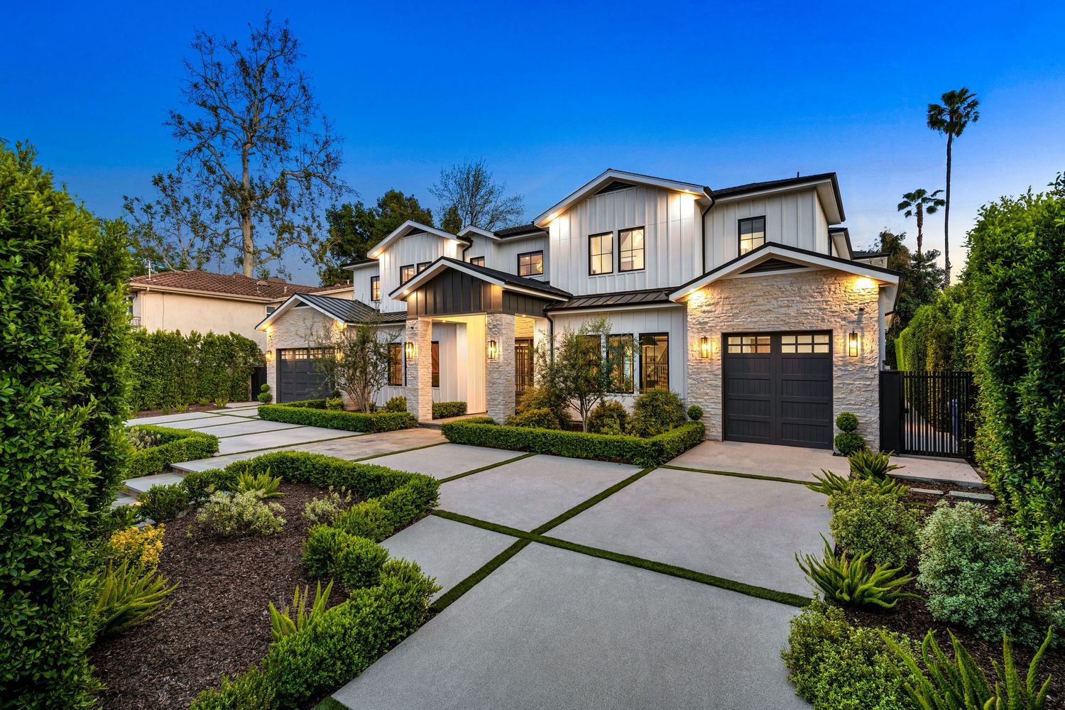 Modern white farmhouse with three-car garage, manicured hedges, and concrete driveway illuminated at dusk.