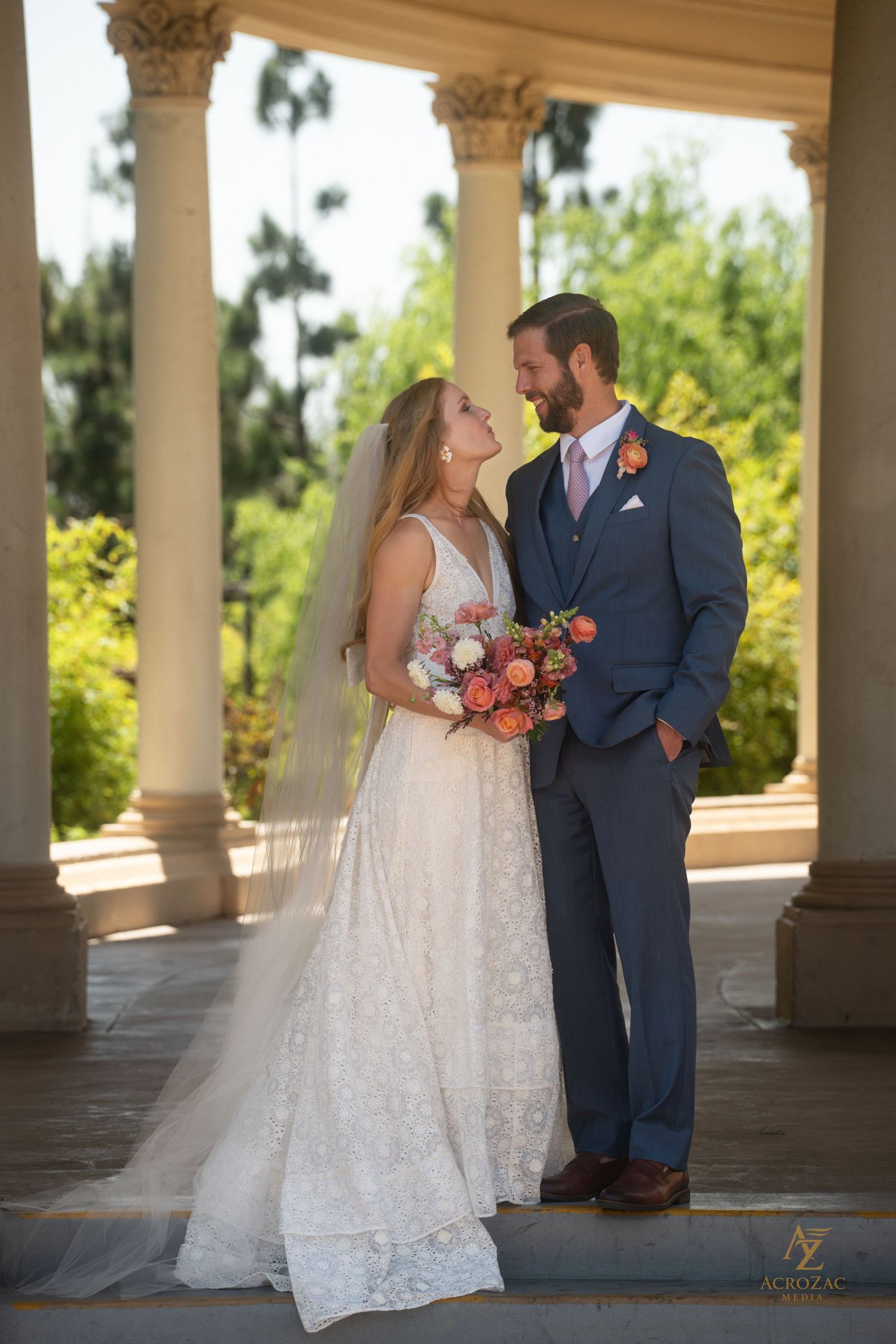 Elegant wedding couple poses under columned archway with garden backdrop.