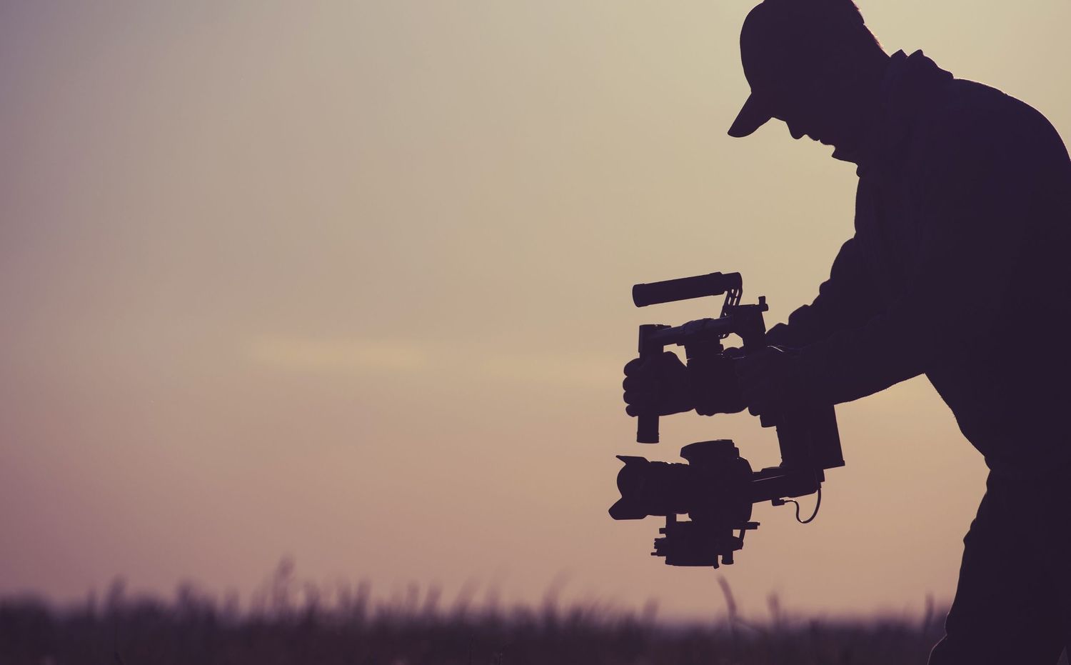 Silhouette of a videographer holding a camera gimbal against a dramatic sunset sky outdoors.