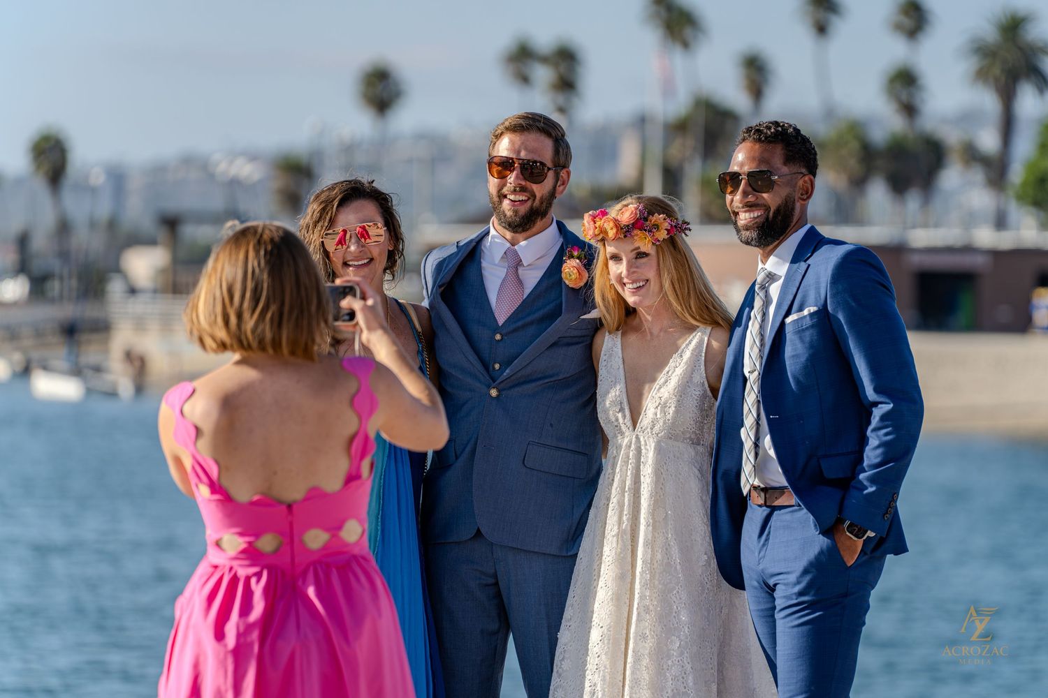 Wedding party laughs together on beach with palm trees in background.