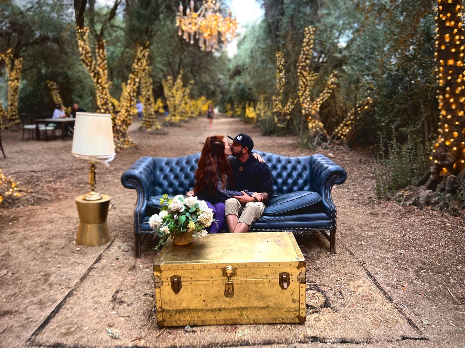 Romantic outdoor setting with blue velvet couch, string lights, and vintage trunk in a garden.