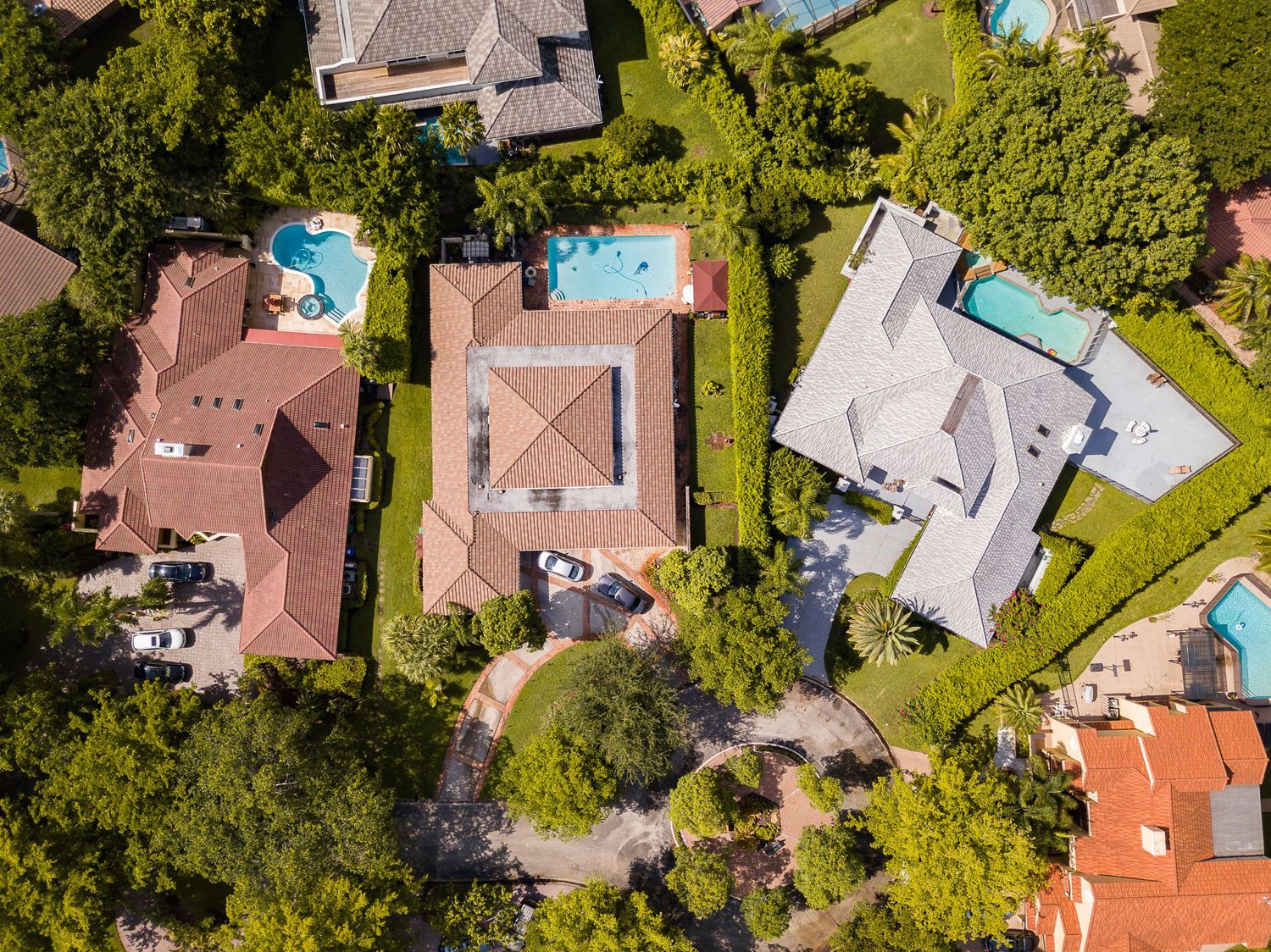 Aerial view of luxury suburban homes with red tile roofs, private pools, and lush green trees in a residential neighborhood.