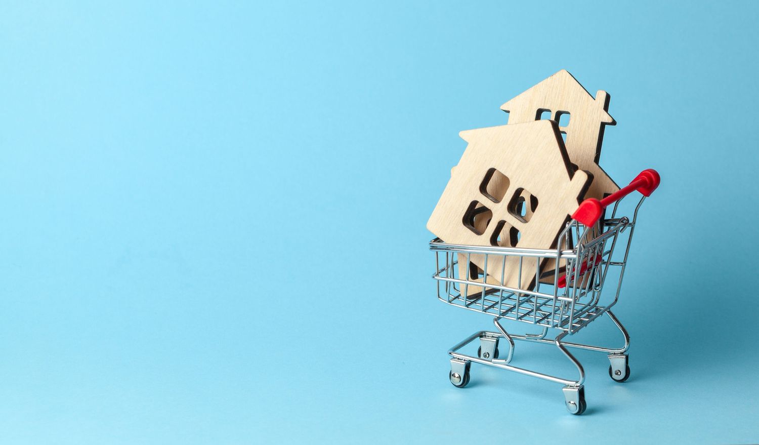 Wooden house models inside a miniature shopping cart on a blue background, symbolizing home buying and real estate.