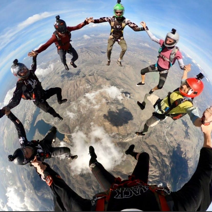 Group of skydivers forming a circular formation during freefall with mountains visible below.