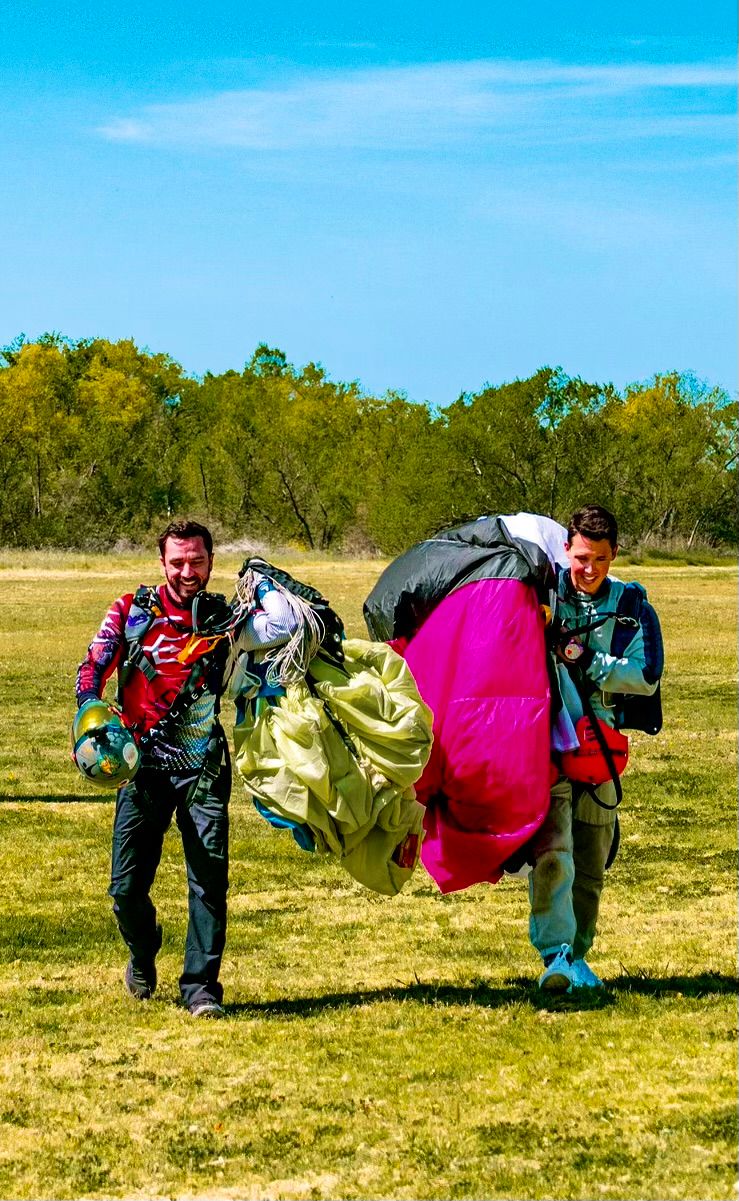 Two skydivers carrying colorful parachute gear walk across a grassy field on a sunny day.