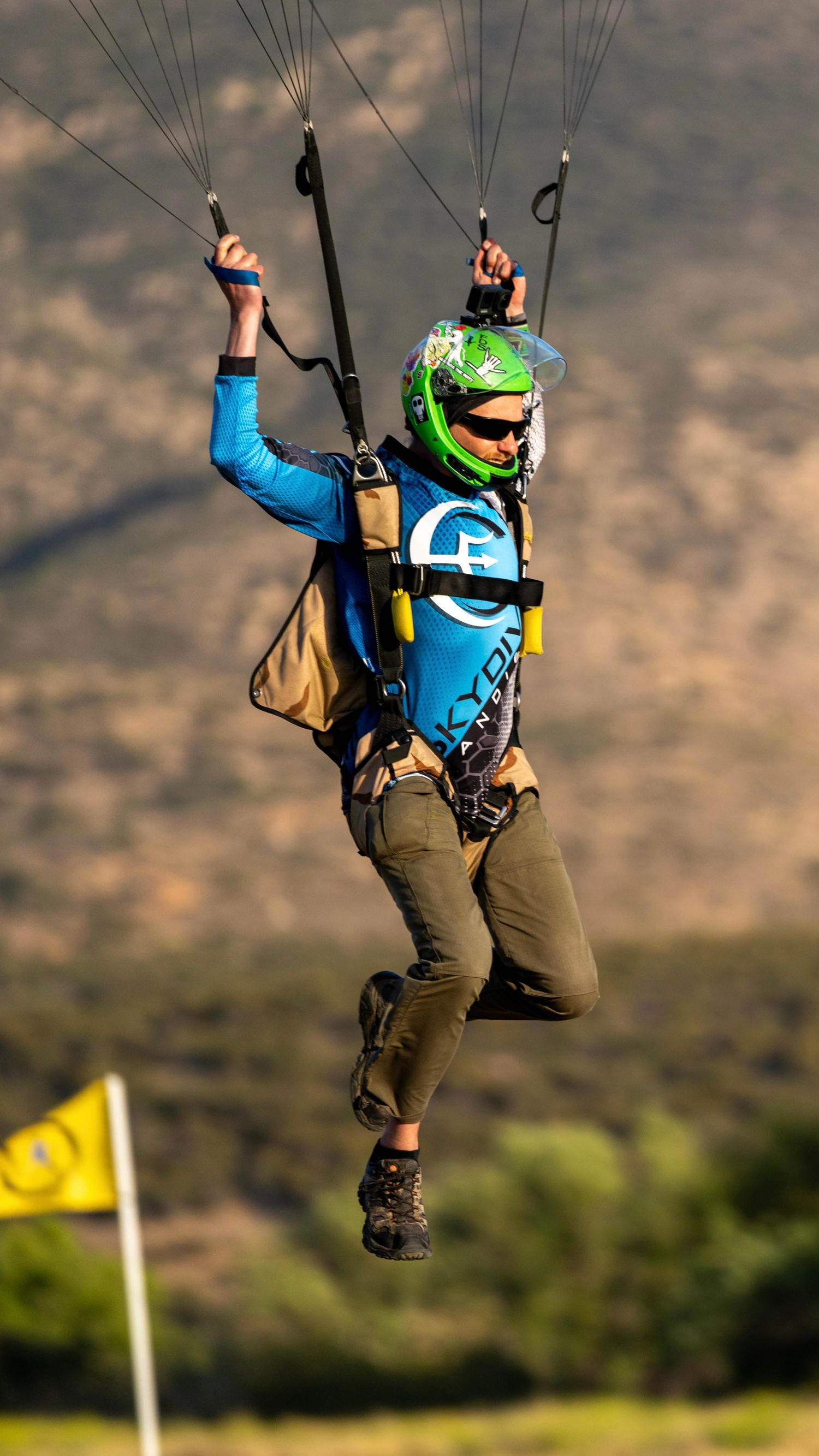 Skydiver in blue suit and green helmet performs controlled descent with parachute lines.