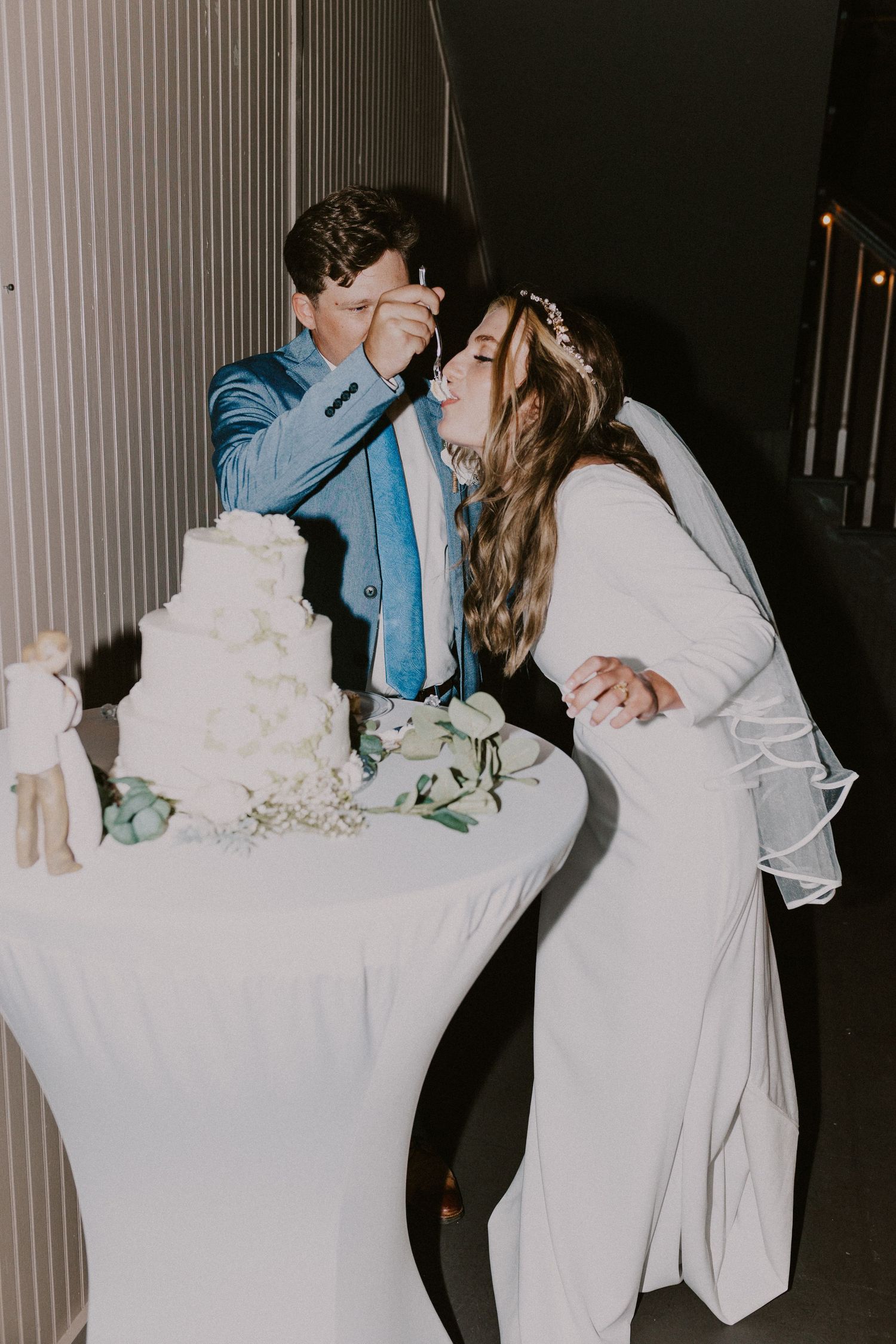 Candid, emotional moment of the bride and groom sharing a piece of their wedding cake during an indoor reception.