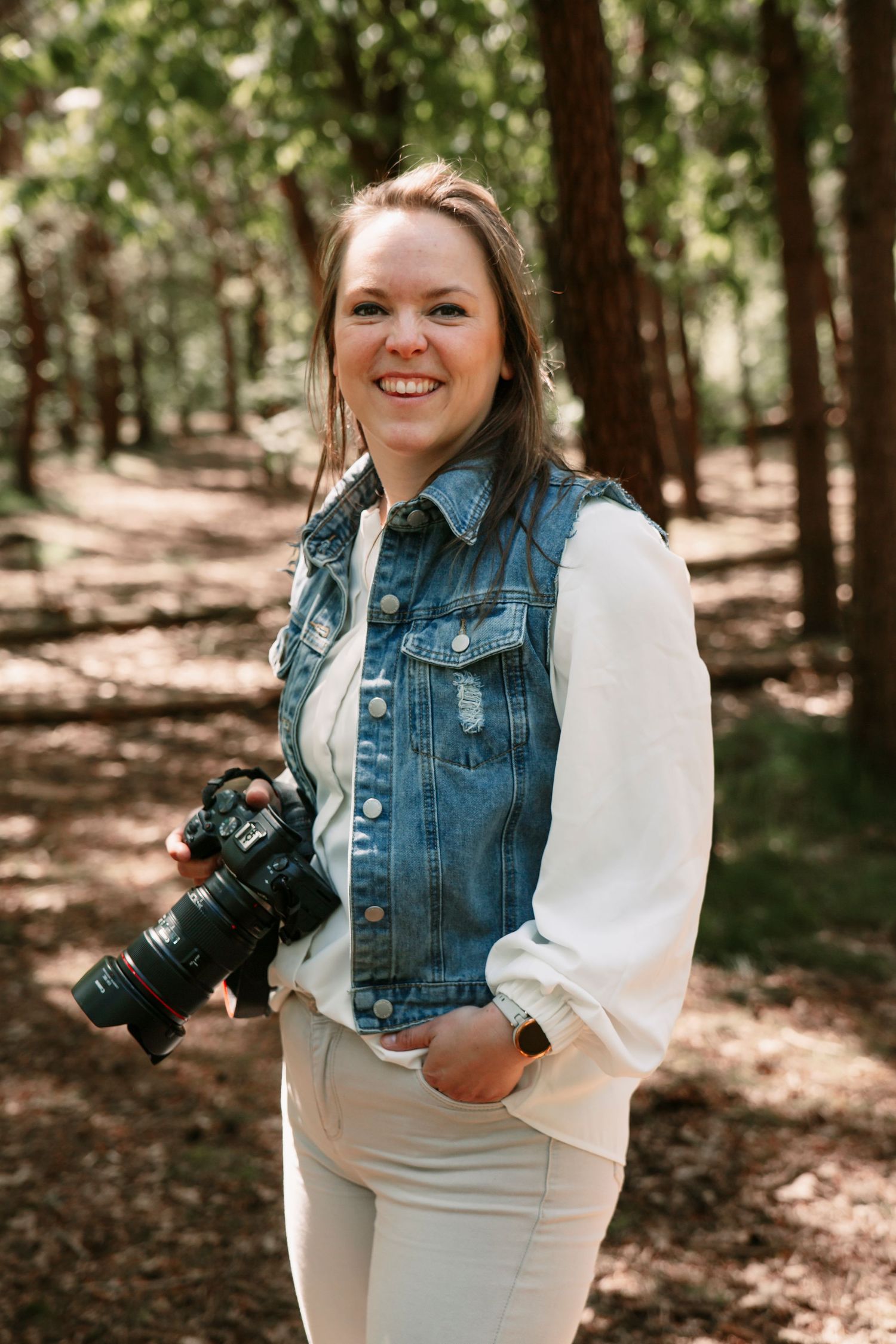 Smiling woman holding a DSLR camera in a forest, wearing a denim vest over a white blouse and light pants.