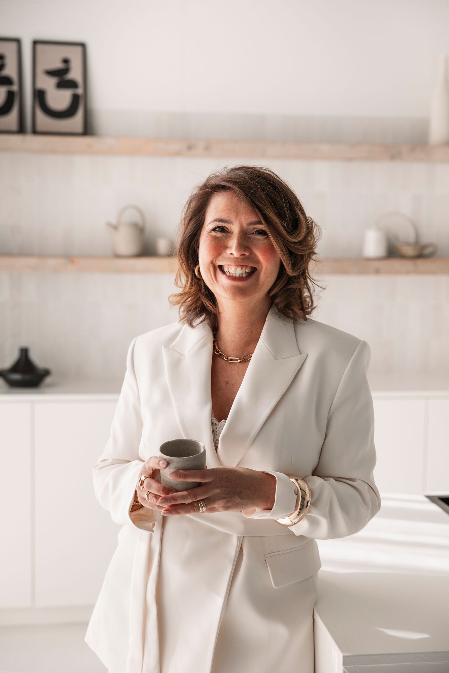 Smiling woman in white blazer holding a ceramic cup in a modern minimalist kitchen with wooden shelves.