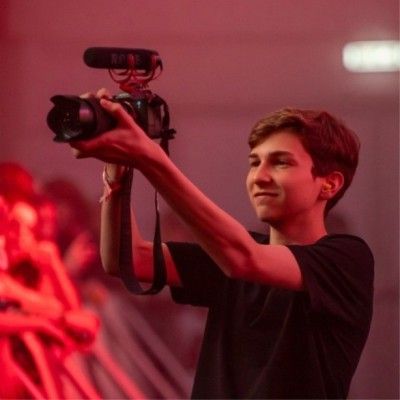 Young videographer holds a professional camera overhead, capturing footage in a red-lit venue while smiling.