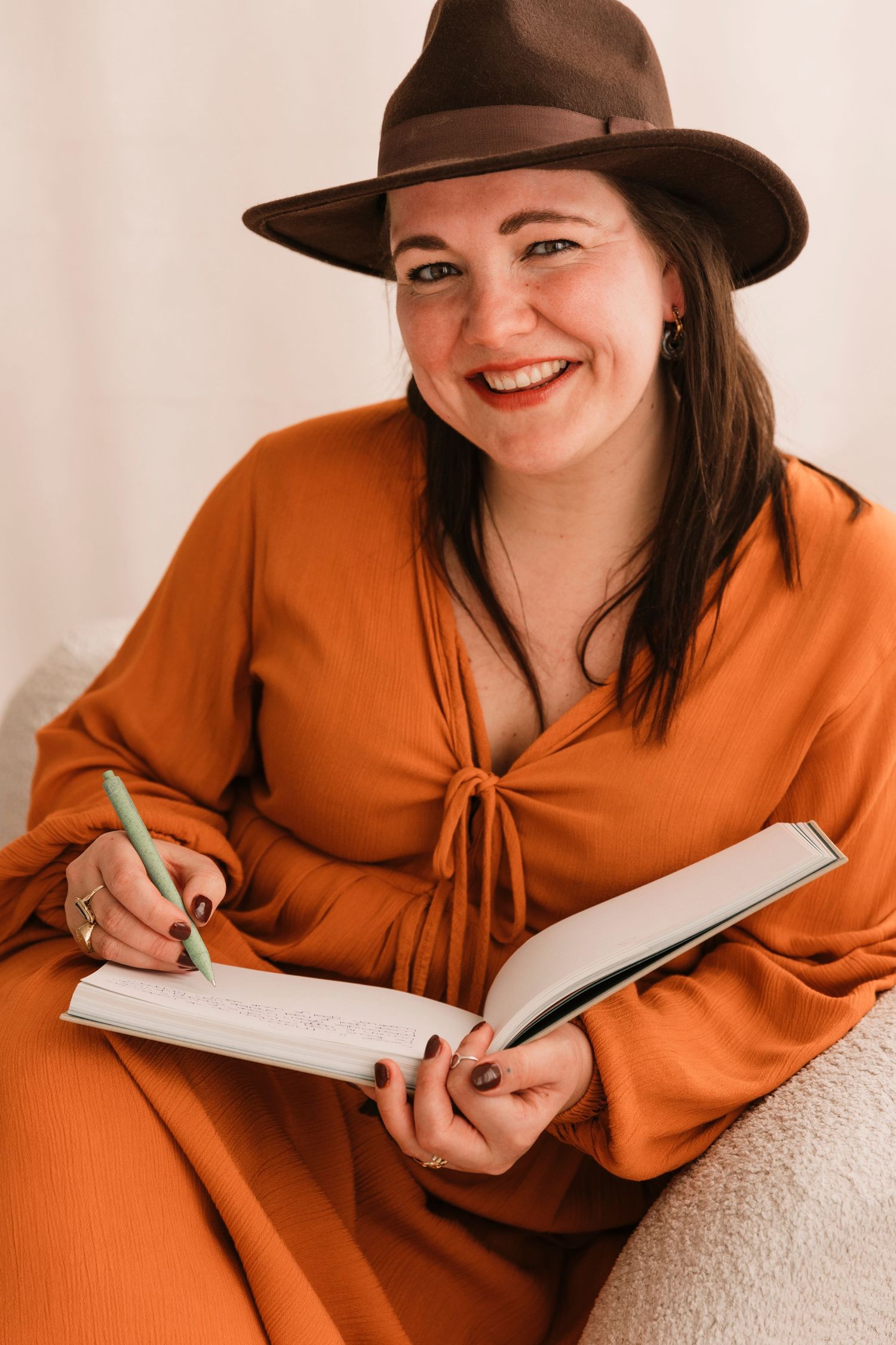 Person in orange top and brown hat writing in notebook while relaxing on couch.