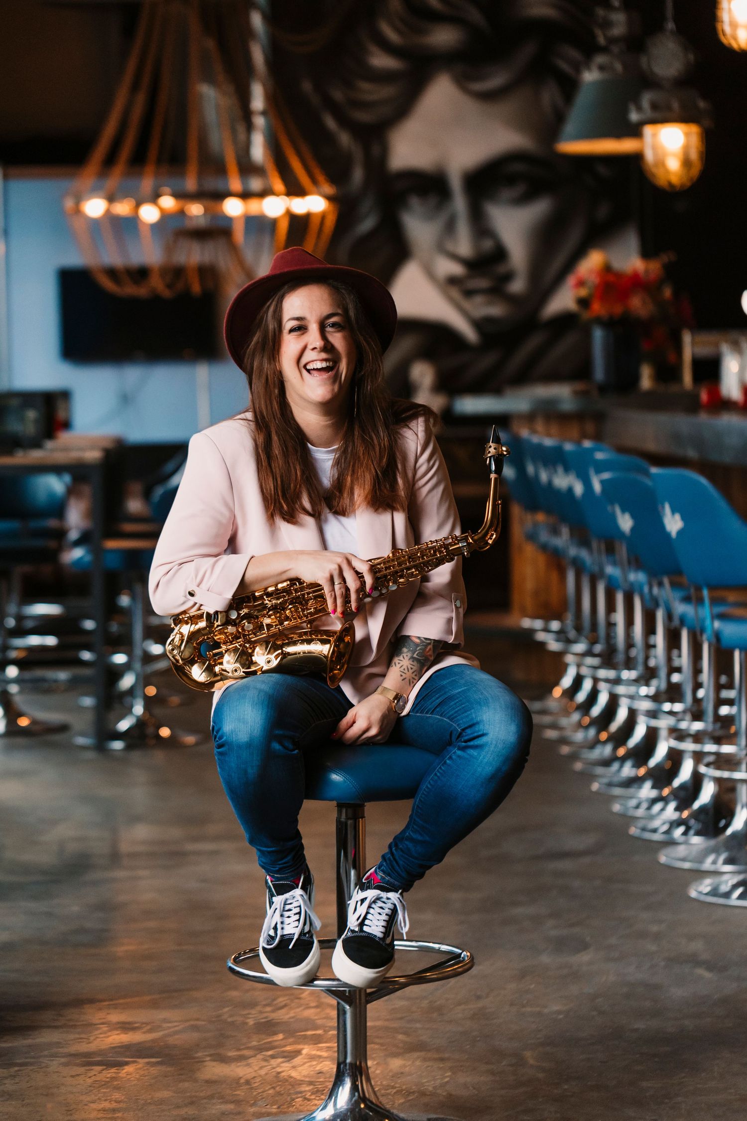 Person in pink sweater holding saxophone while sitting on bar stool in dimly lit venue.