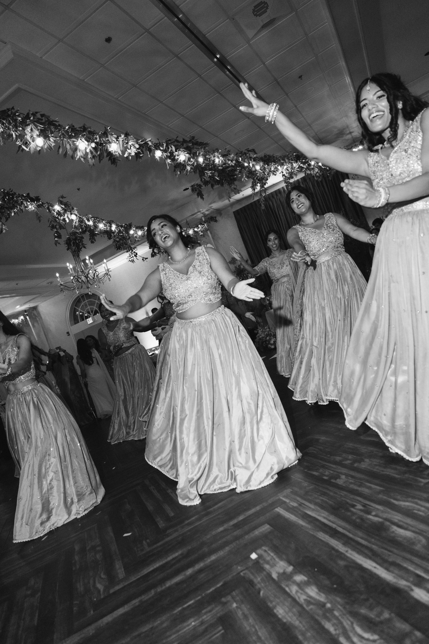 Bridesmaids in elegant gowns dancing joyfully at a wedding reception in black and white photo.