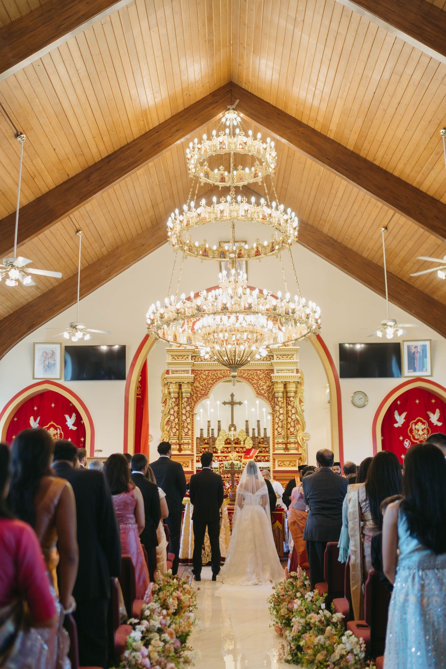 Wedding ceremony inside ornate church with crystal chandeliers as couple walks down floral aisle.
