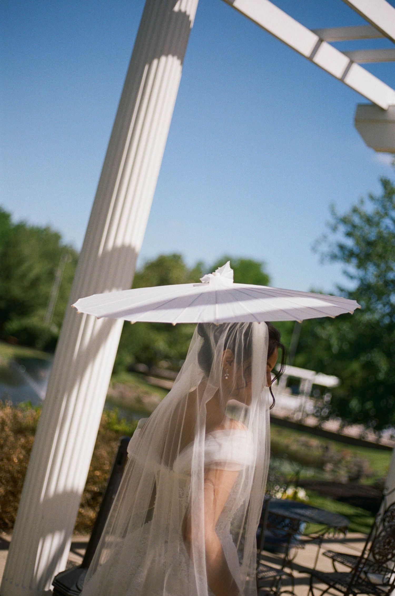 Bride holding white parasol with veil flowing under pergola columns on a bright sunny day.