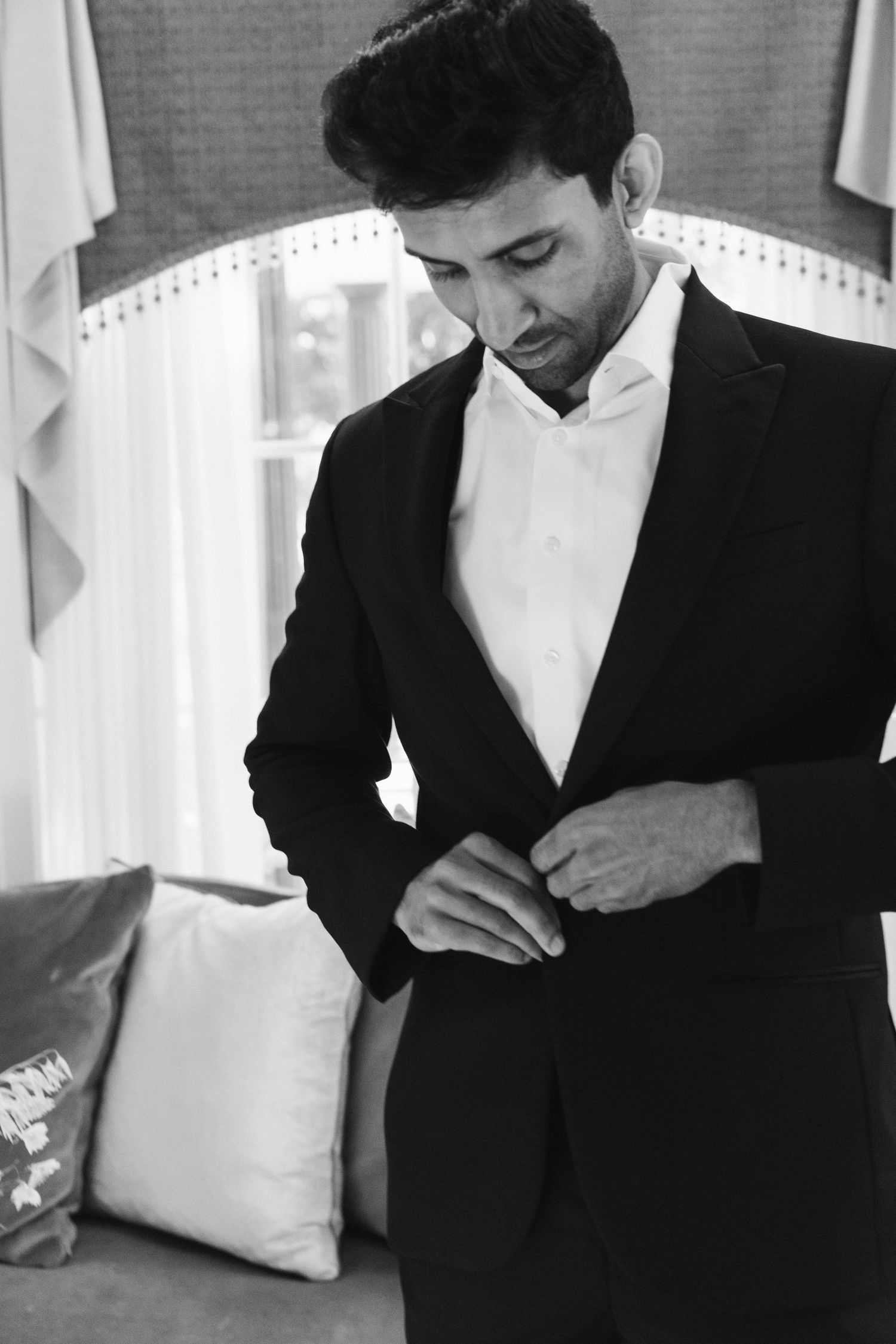 A groom in a black tuxedo buttons his jacket while preparing for his wedding day.