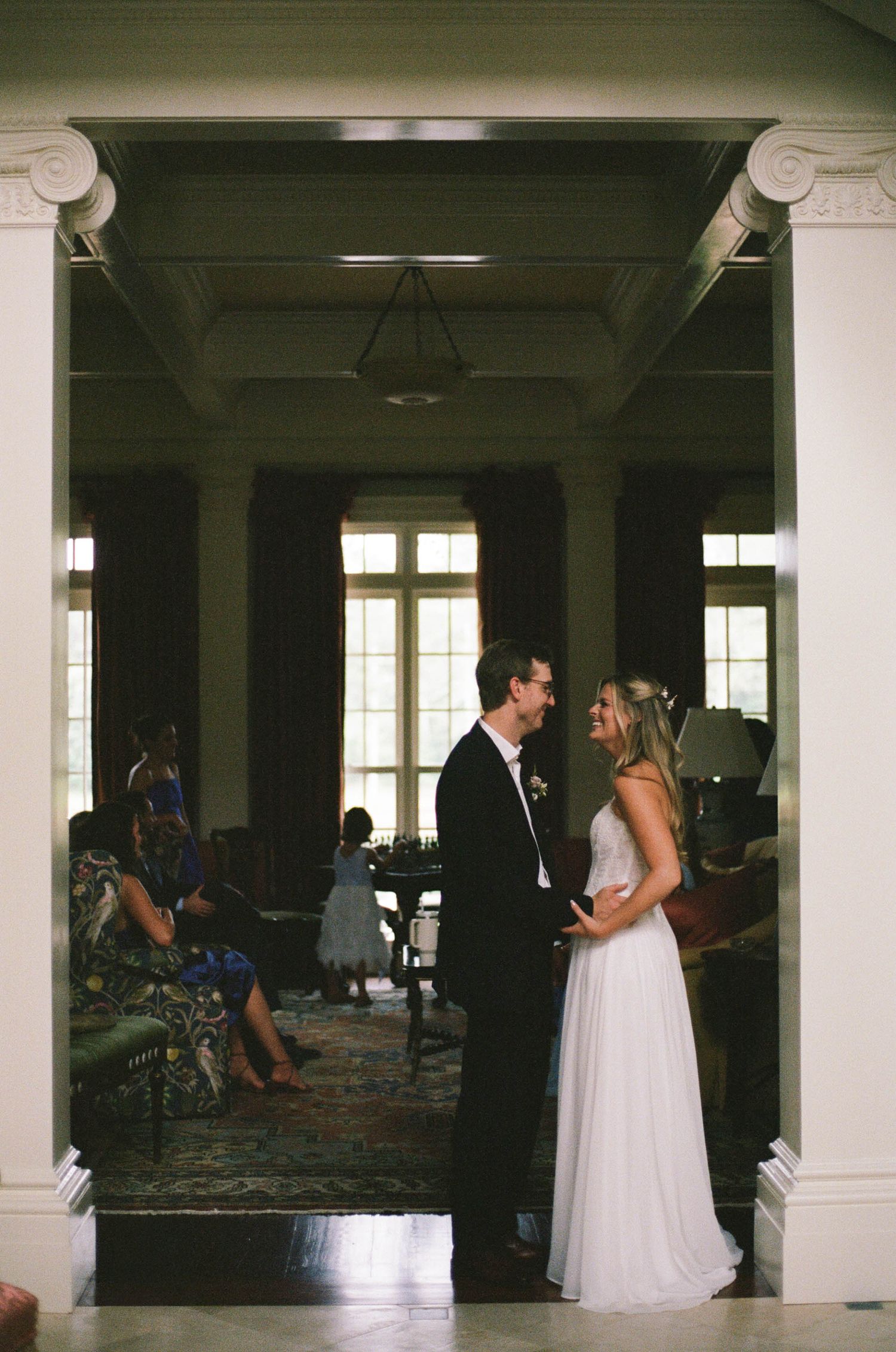 Bride and groom share intimate moment framed by white columns in elegant historic mansion doorway.