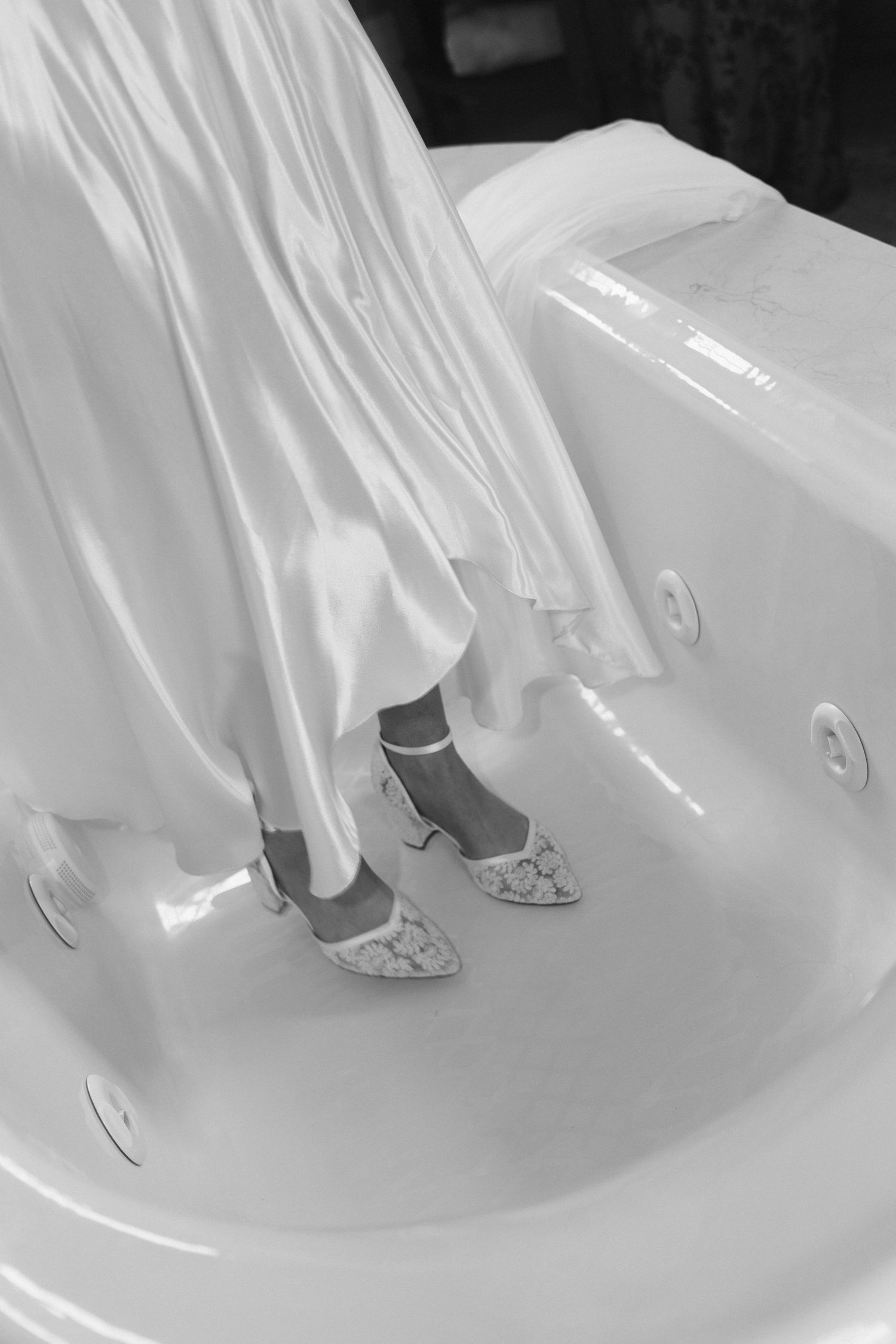 Black and white photo of bride in white gown standing in bathtub wearing heeled shoes.