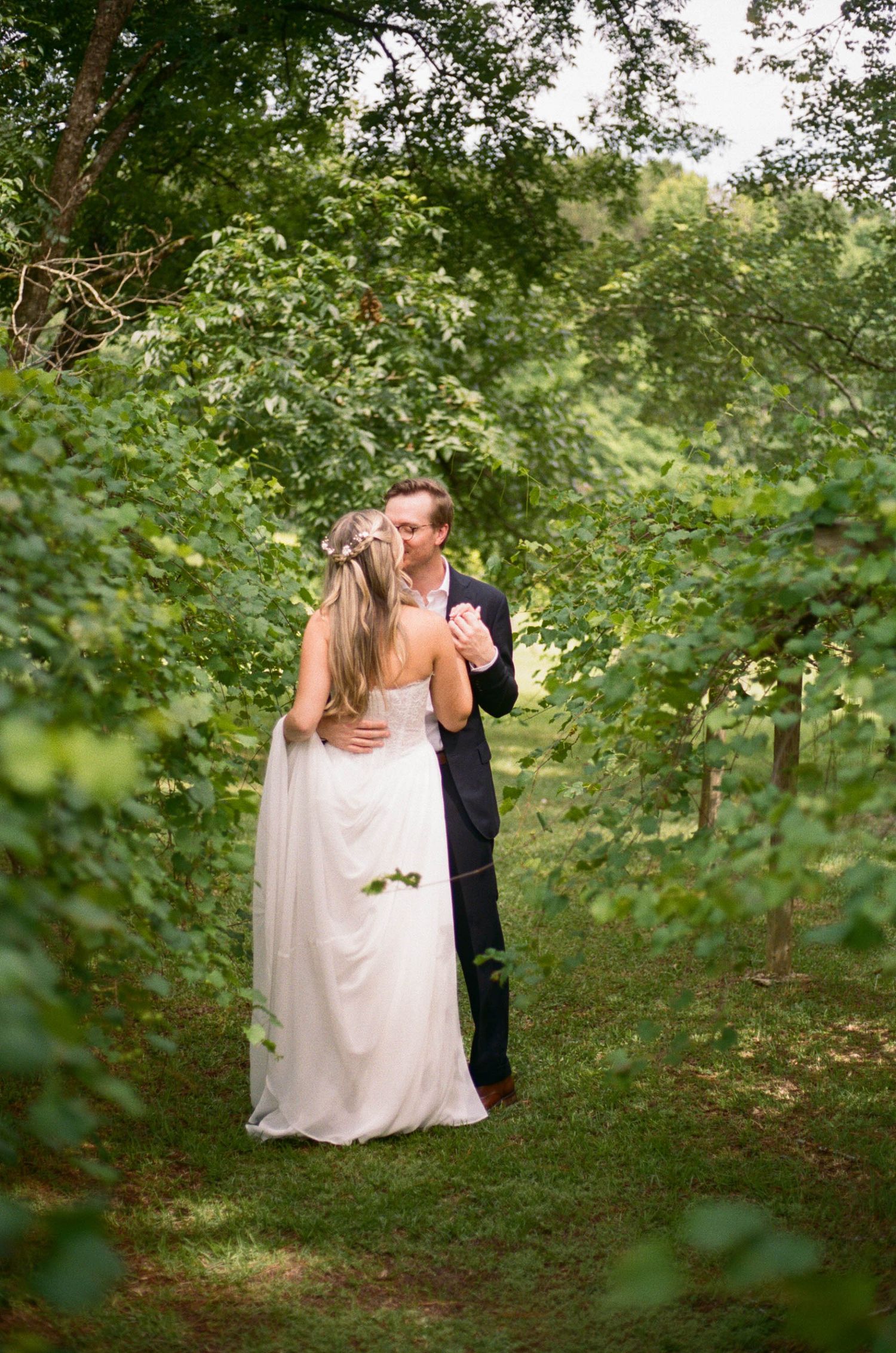 Bride and groom share a romantic kiss along a lush green tree-lined garden path on their wedding day.