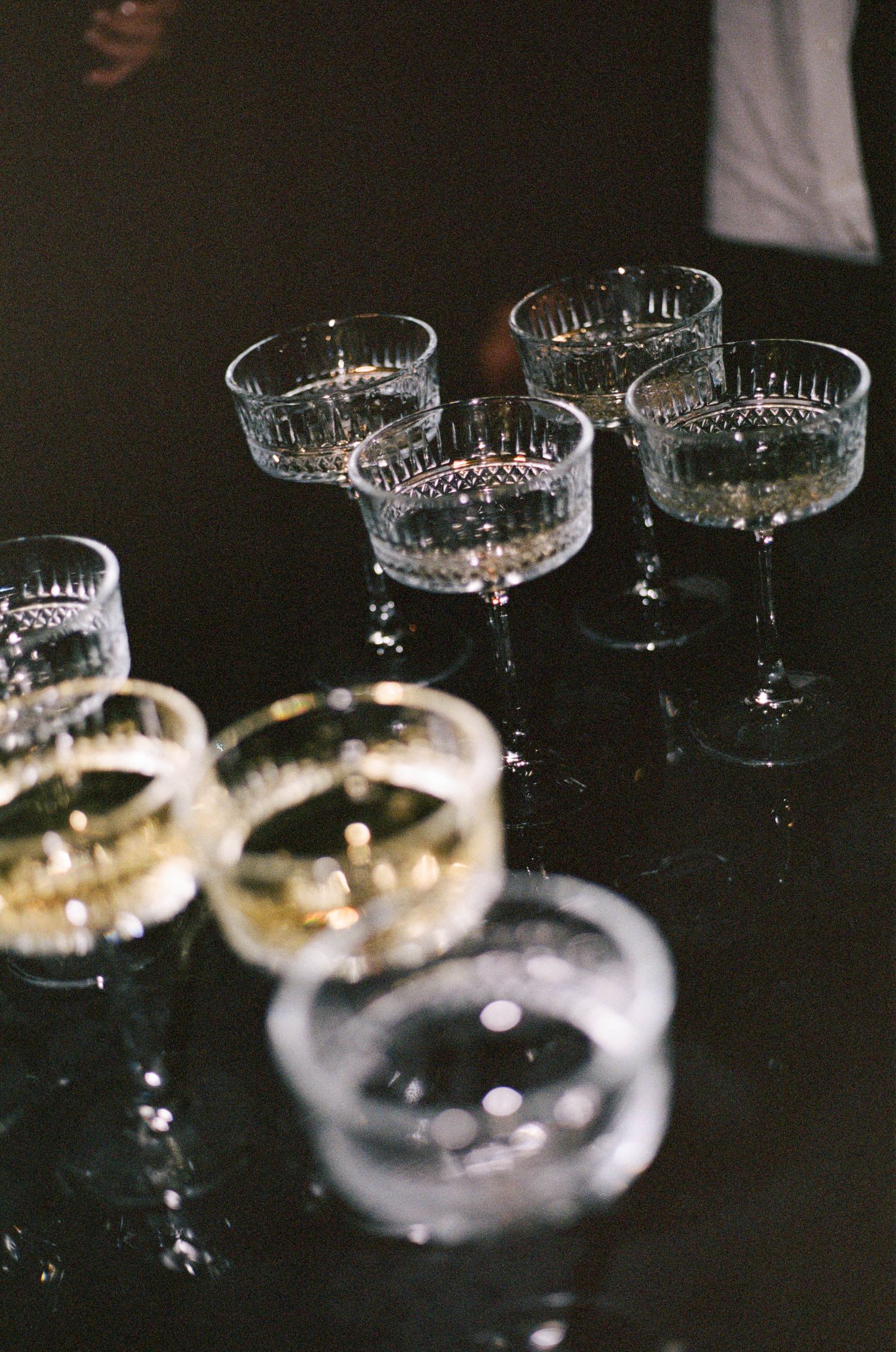 Elegant crystal coupe glasses arranged on a dark surface reflecting ambient light.