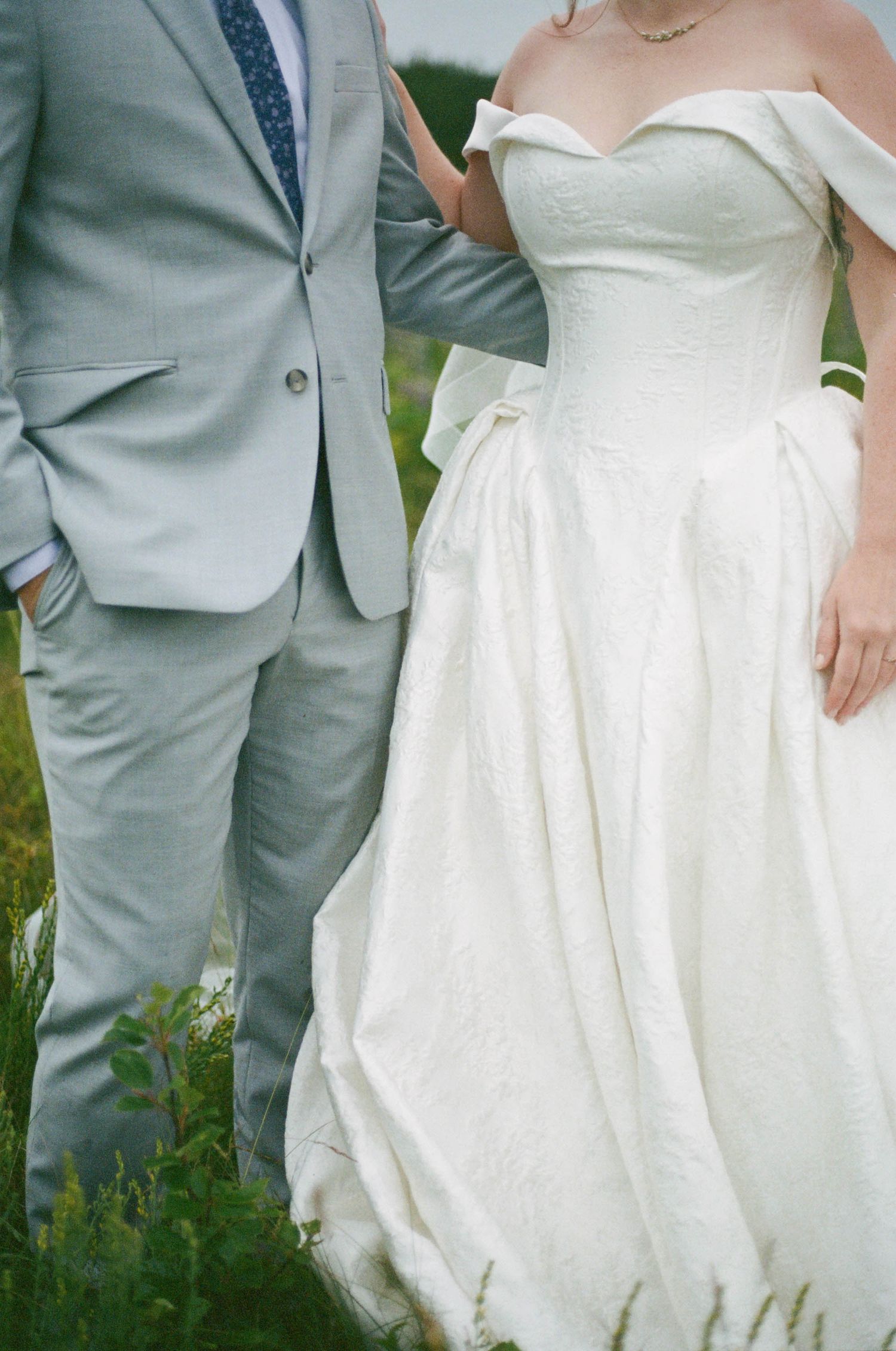Detail shot of a bride in white dress standing next to groom in light grey suit among wildflowers.