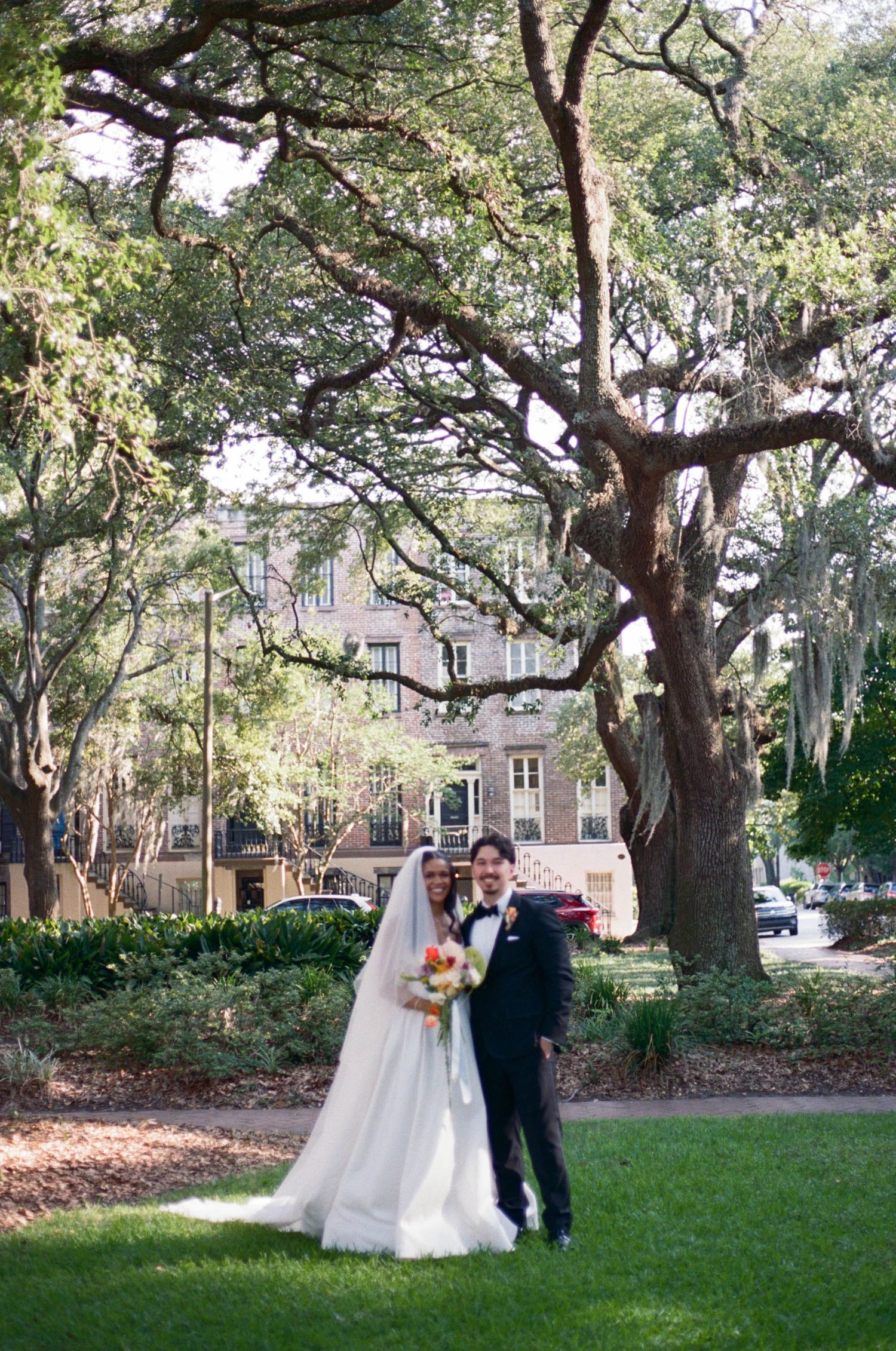 A wedding couple poses for photos under majestic oak trees in a garden setting.