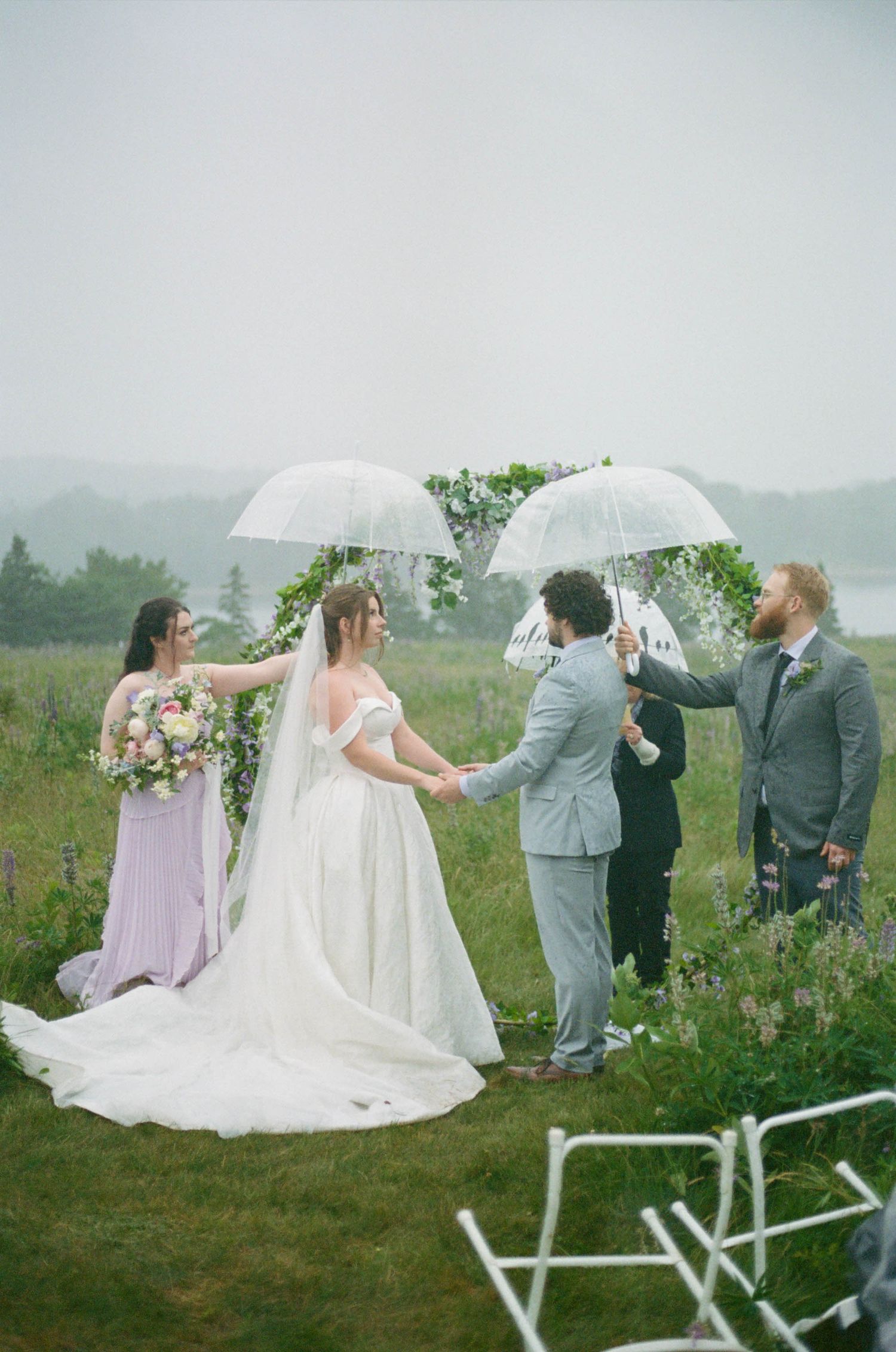 Wedding party poses with umbrellas in a misty field during light rain.