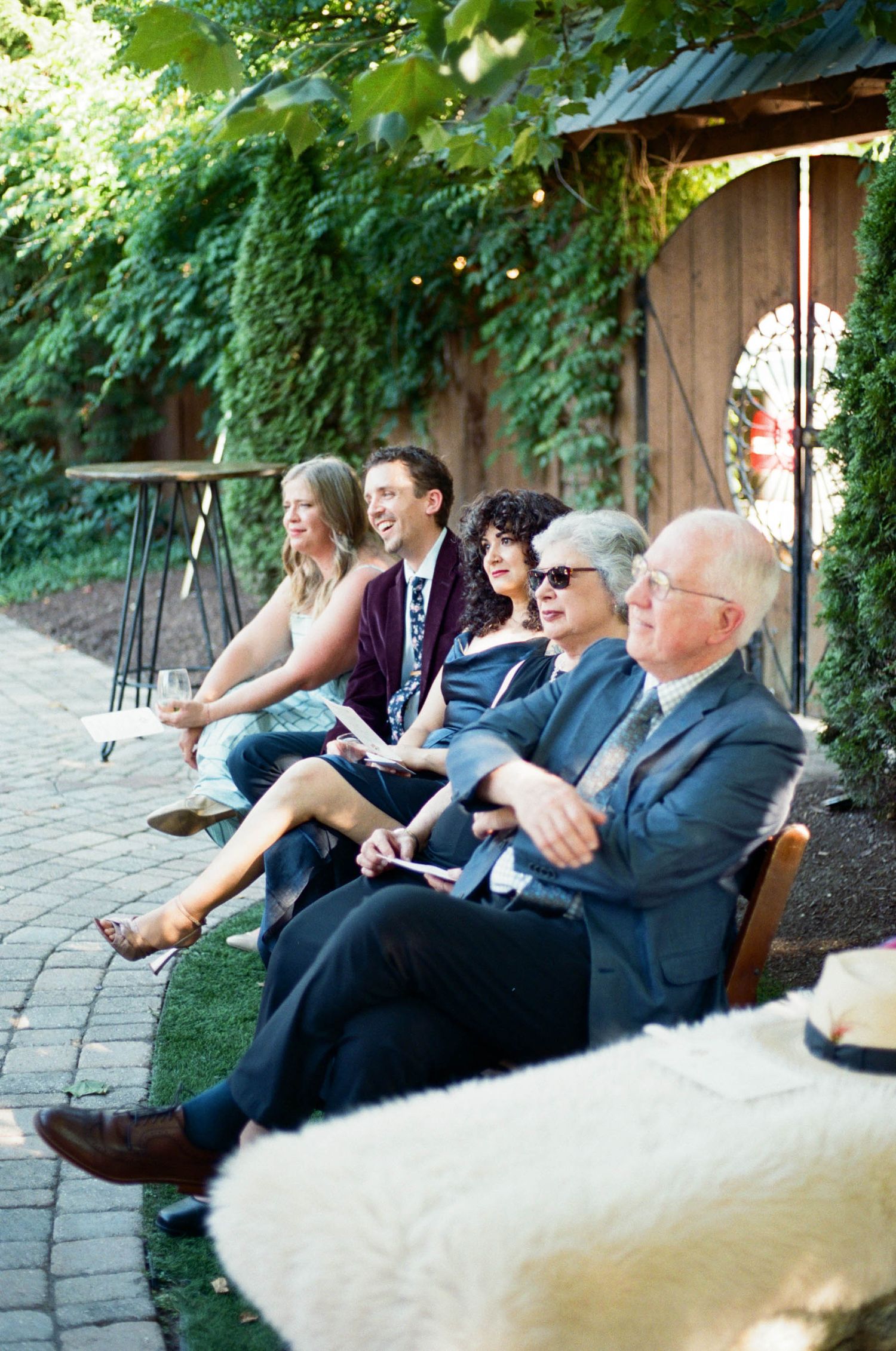 Wedding guests seated on benches laugh and smile during an outdoor ceremony.