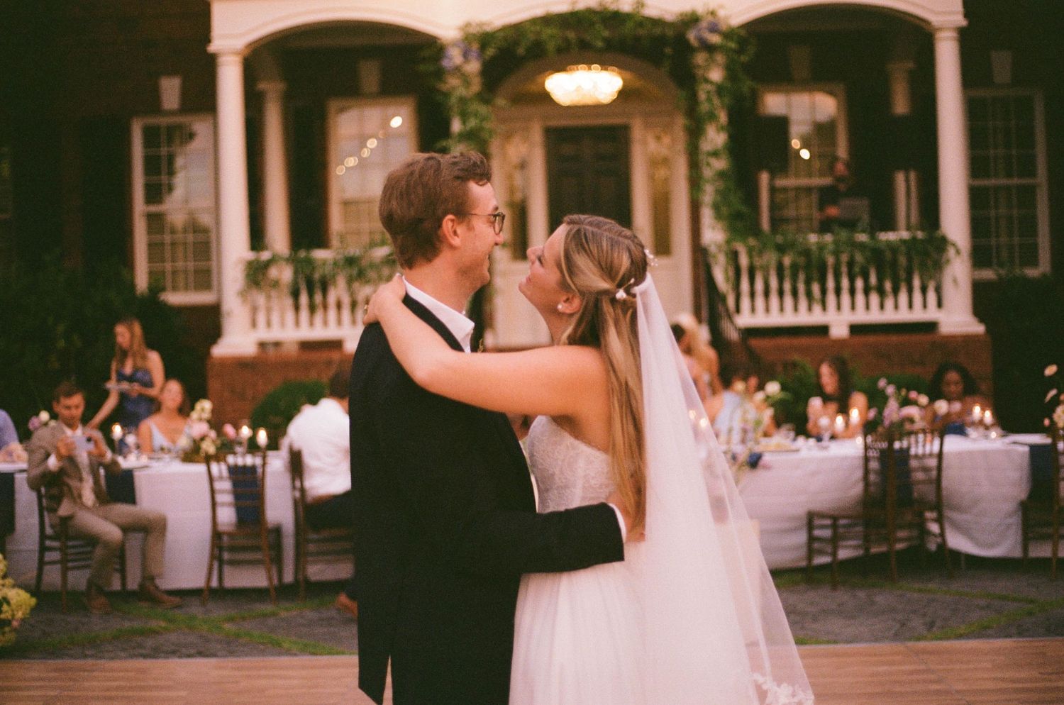 A bride and groom share their first dance outdoors at an elegant evening wedding reception with guests seated at candlelit tables.