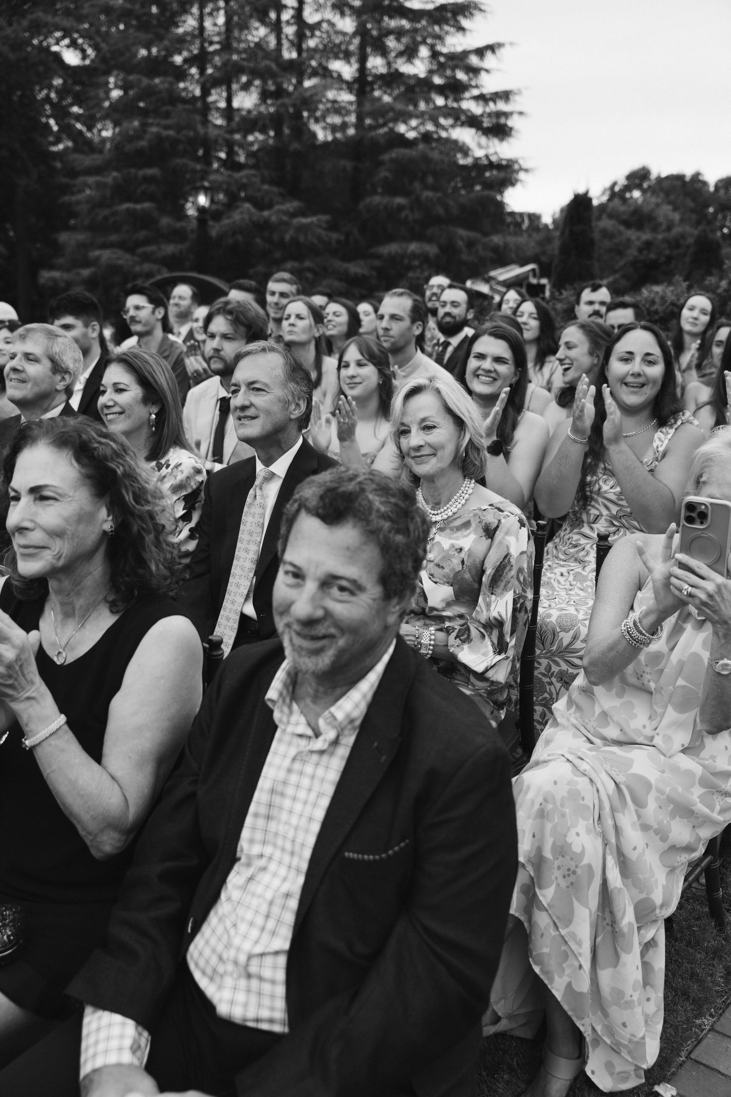 Black and white photo of smiling wedding guests seated outdoors at a garden ceremony.