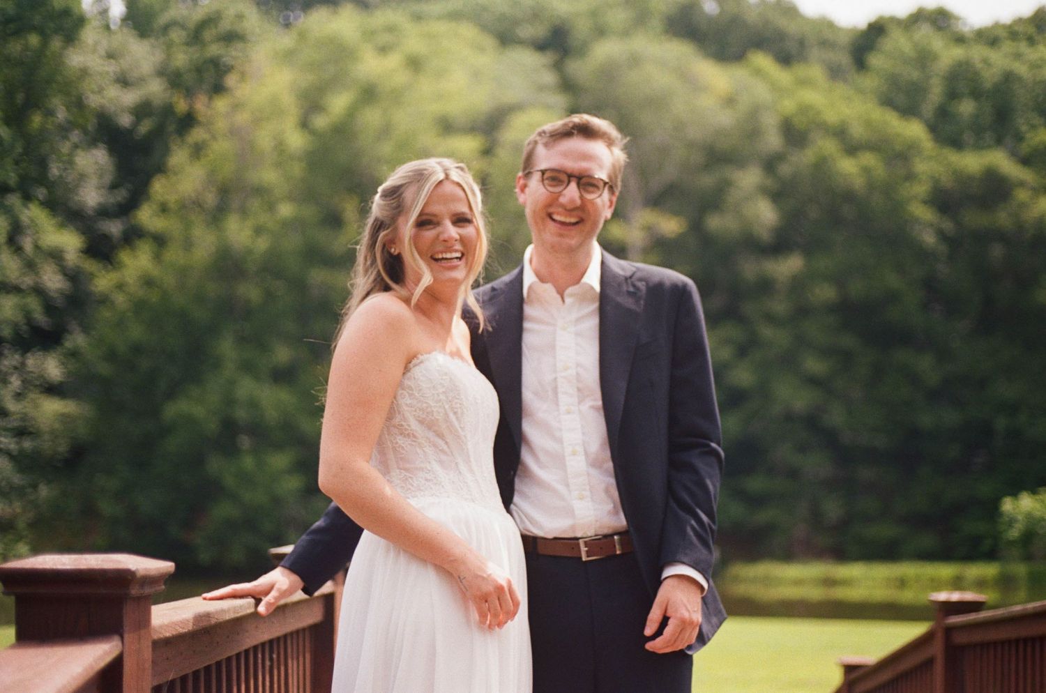Happy bride and groom smile together on a wooden bridge surrounded by lush green trees.