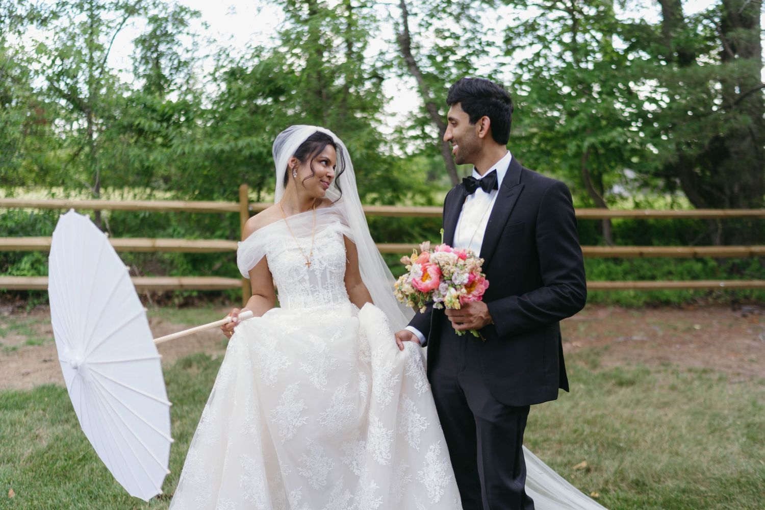 Bride holding a white parasol and bouquet stands with groom in tuxedo outdoors at wedding.