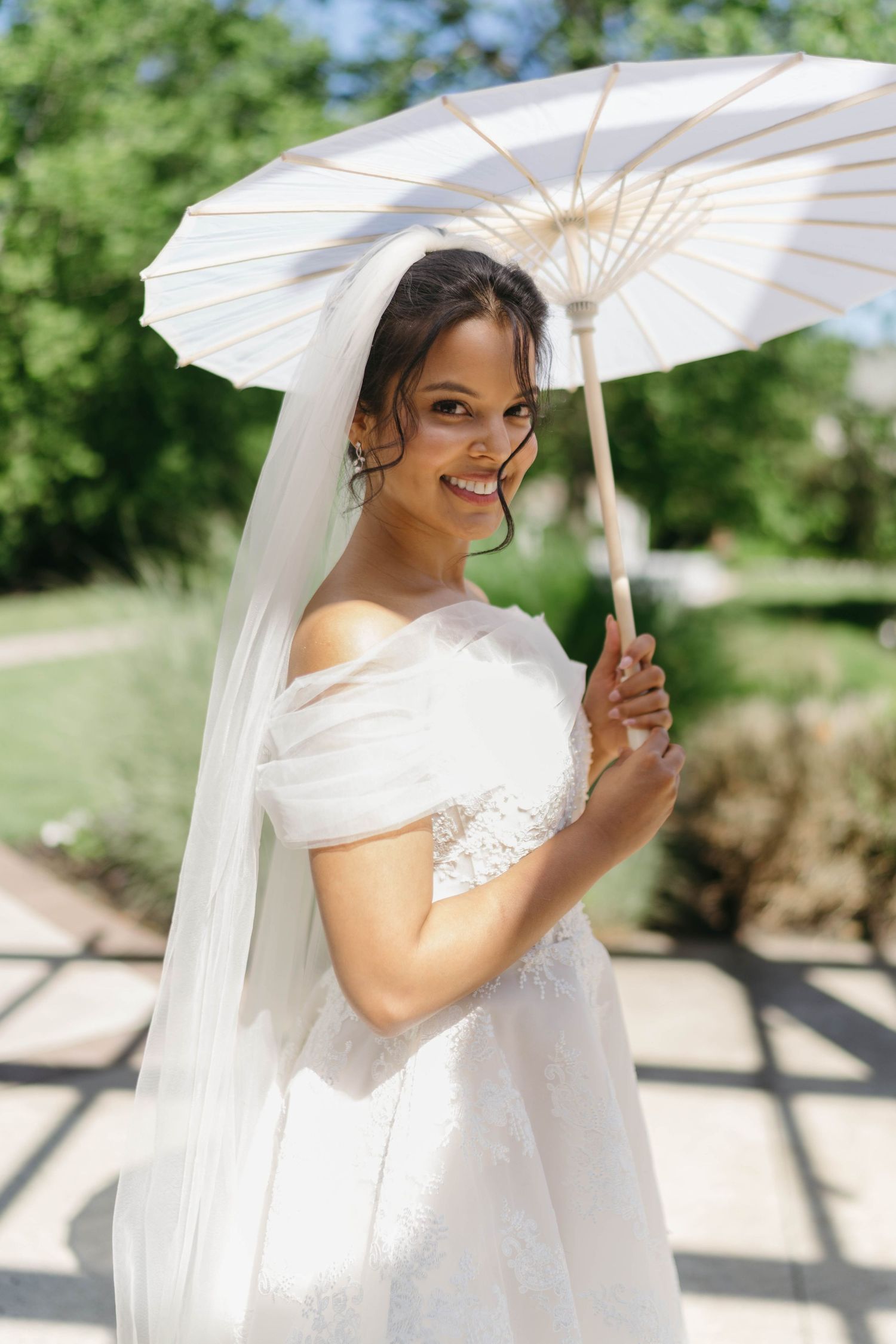 A smiling bride in a white off-shoulder gown holds a parasol umbrella outdoors on her wedding day.