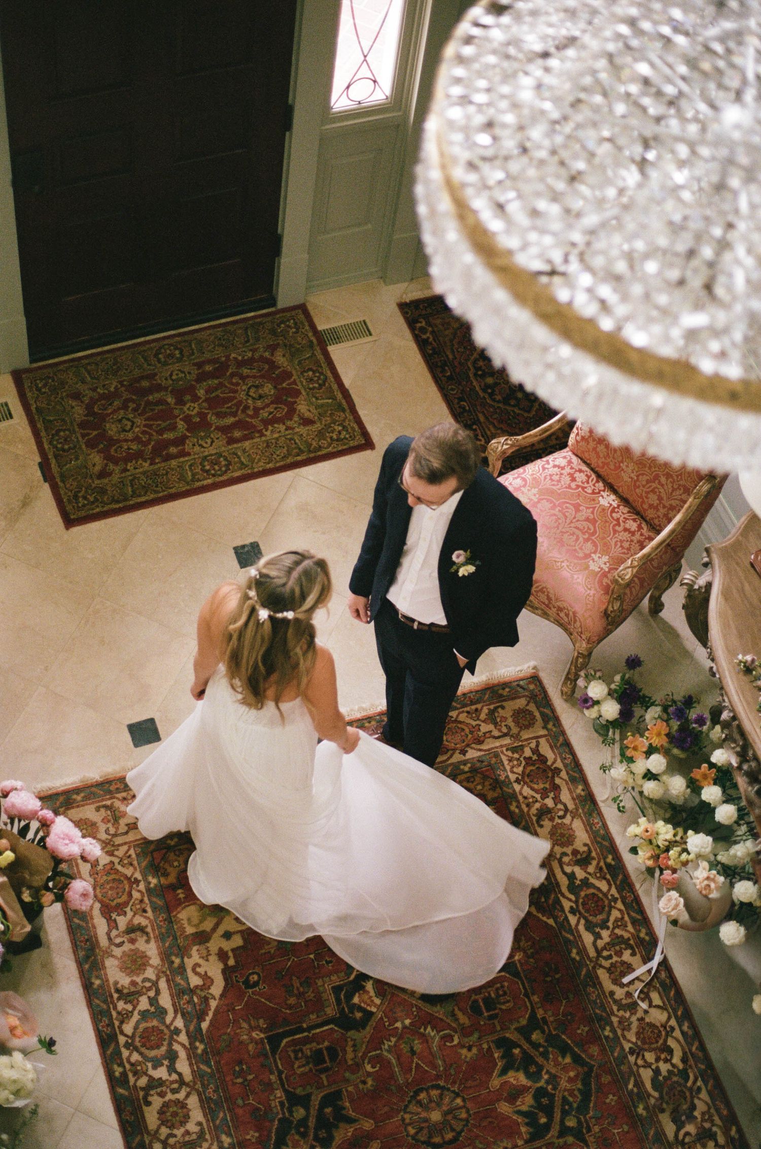 Aerial view of bride and groom sharing a first look moment in an elegant foyer with crystal chandelier and ornate rug.