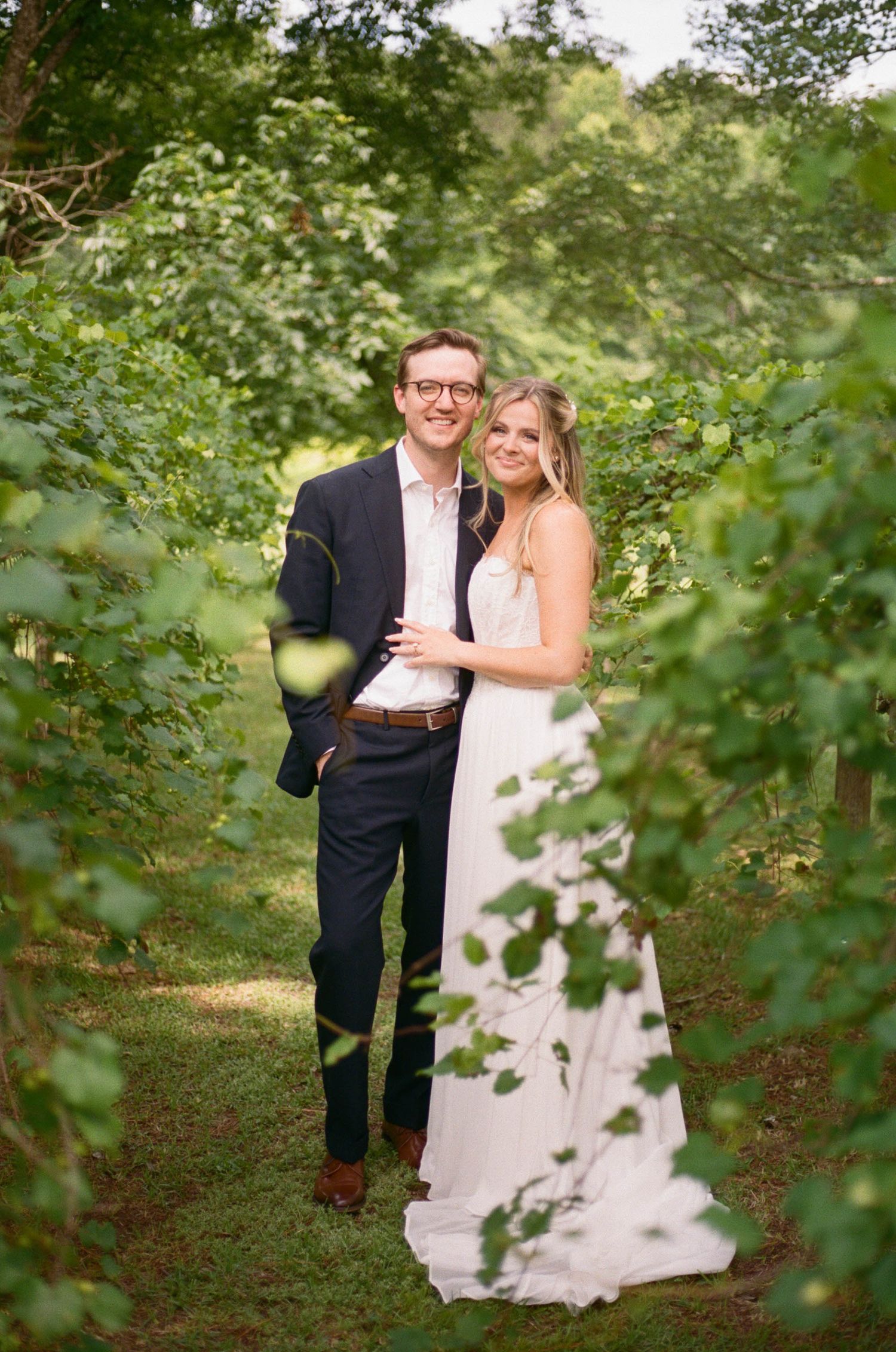 Smiling bride and groom pose together in a beautiful green vineyard pathway on their wedding day.