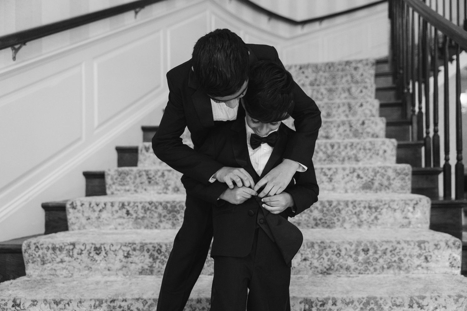 Two men in black tuxedos adjusting formal wear on elegant staircase in black and white wedding photo.