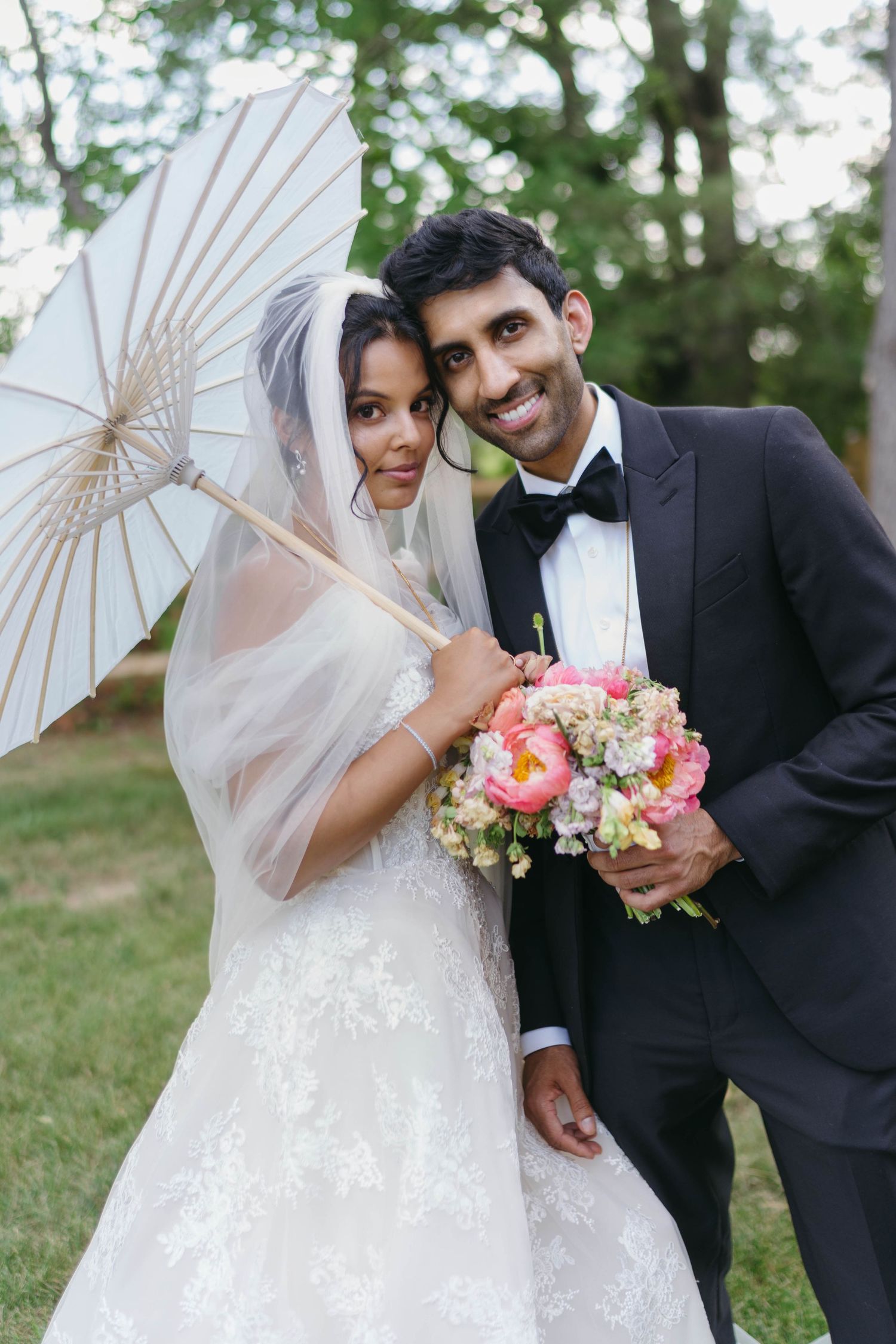 Bride and groom posing outdoors, bride holding white parasol and veil, groom in tuxedo holding pink bouquet.