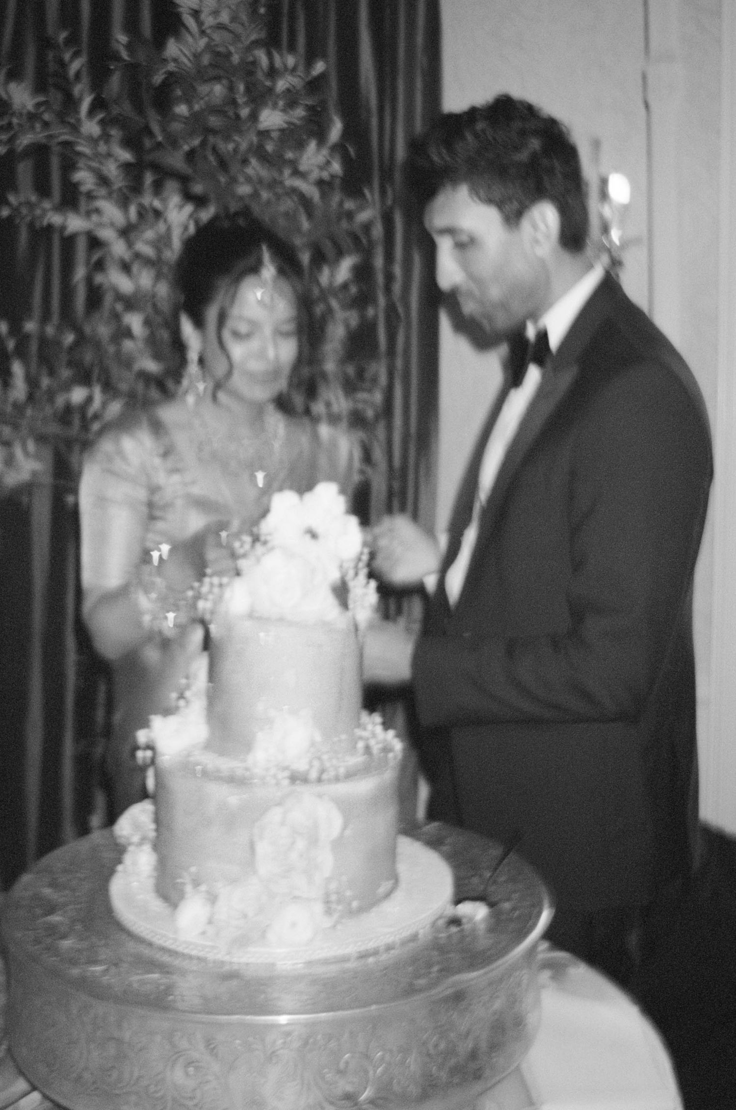 Black and white photo of bride and groom cutting tiered wedding cake decorated with white floral topper.