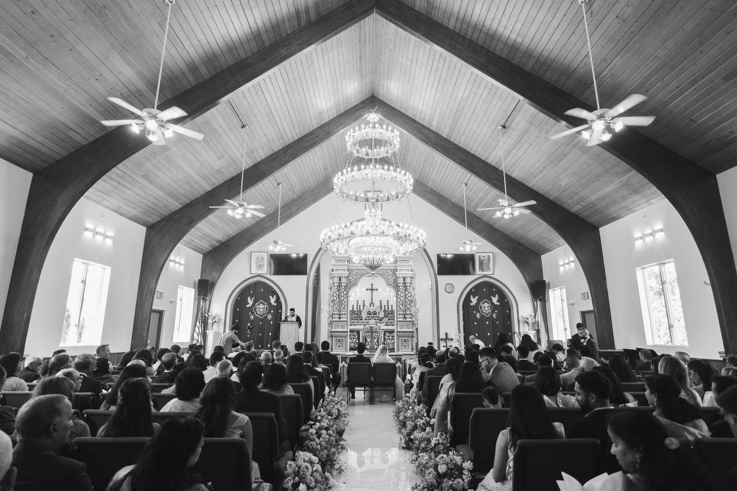 Wide-angle black and white photo of packed church ceremony with chandelier lighting and floral altar decorations.