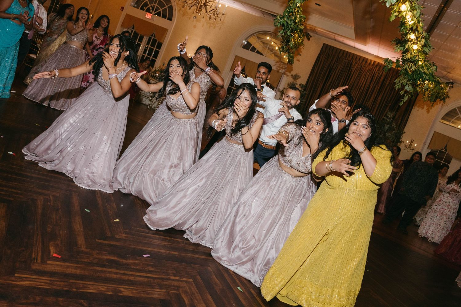 Bridesmaids in silver gowns dance joyfully on the wedding reception floor with guests in elegant Indian attire.