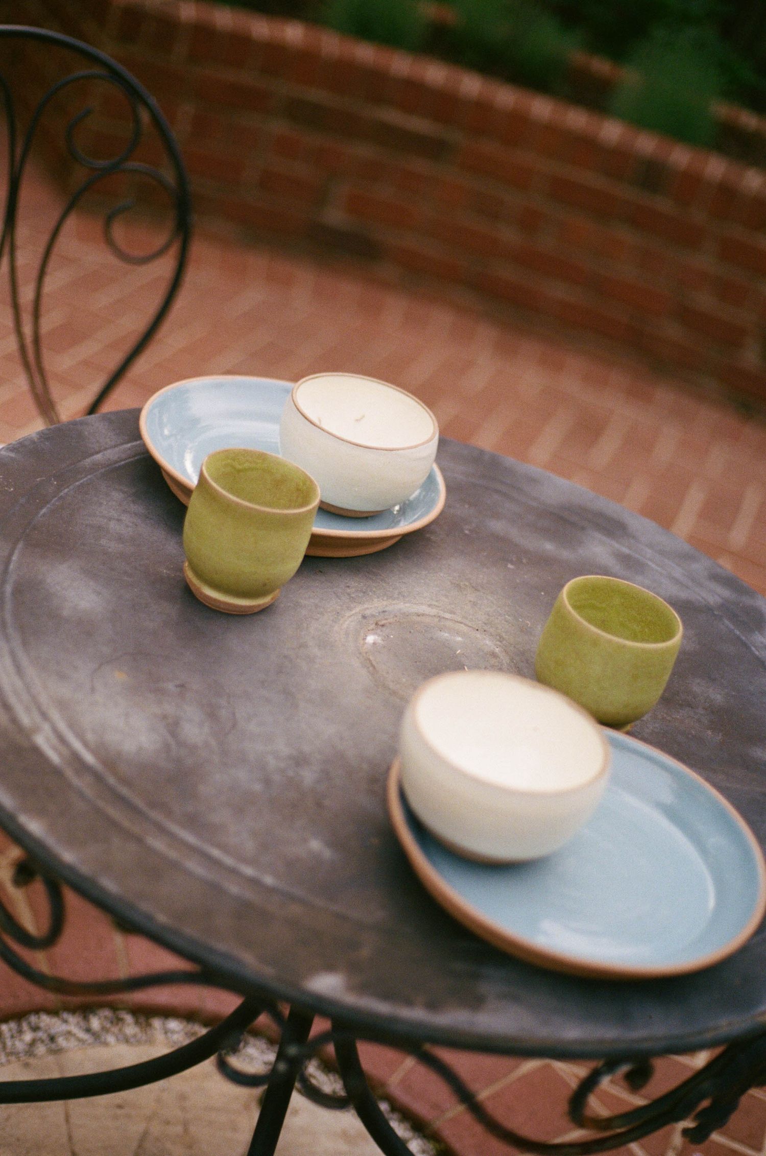 Handmade ceramic bowls and green cups on a rustic metal outdoor table with brick wall background.