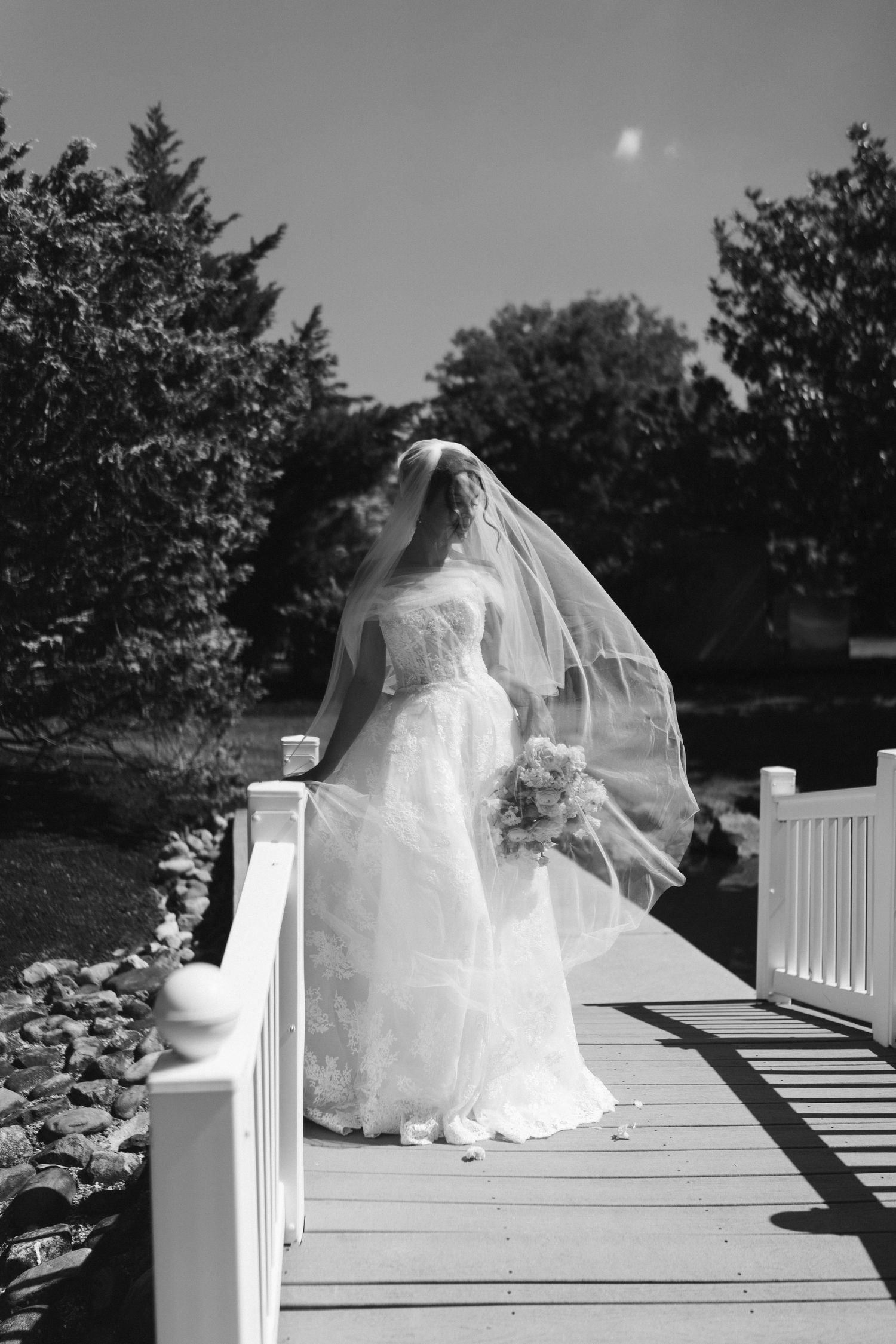 Bride in white lace wedding gown with flowing veil standing on wooden bridge surrounded by lush greenery.