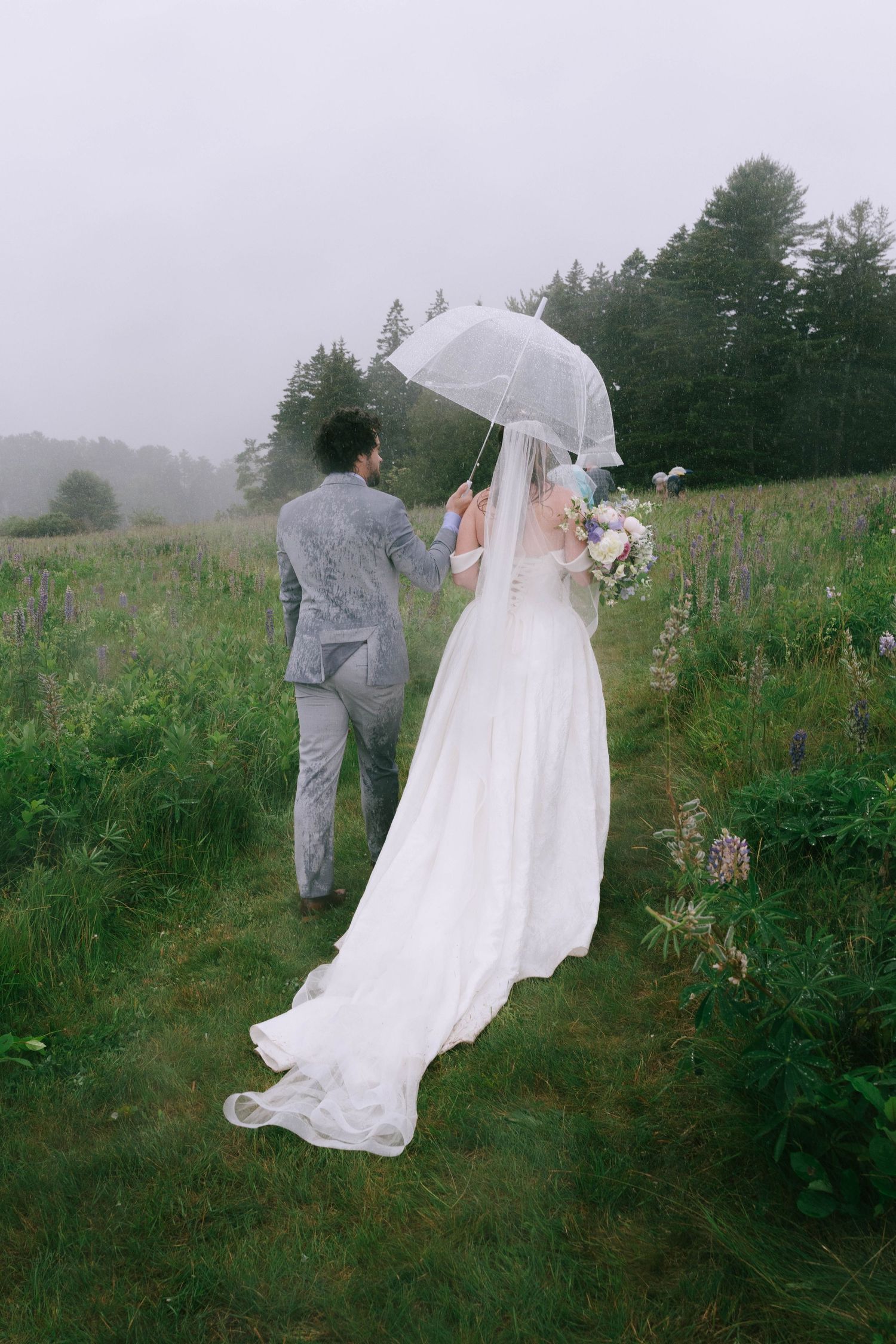 A couple walks through a misty field holding a clear umbrella during their wedding.