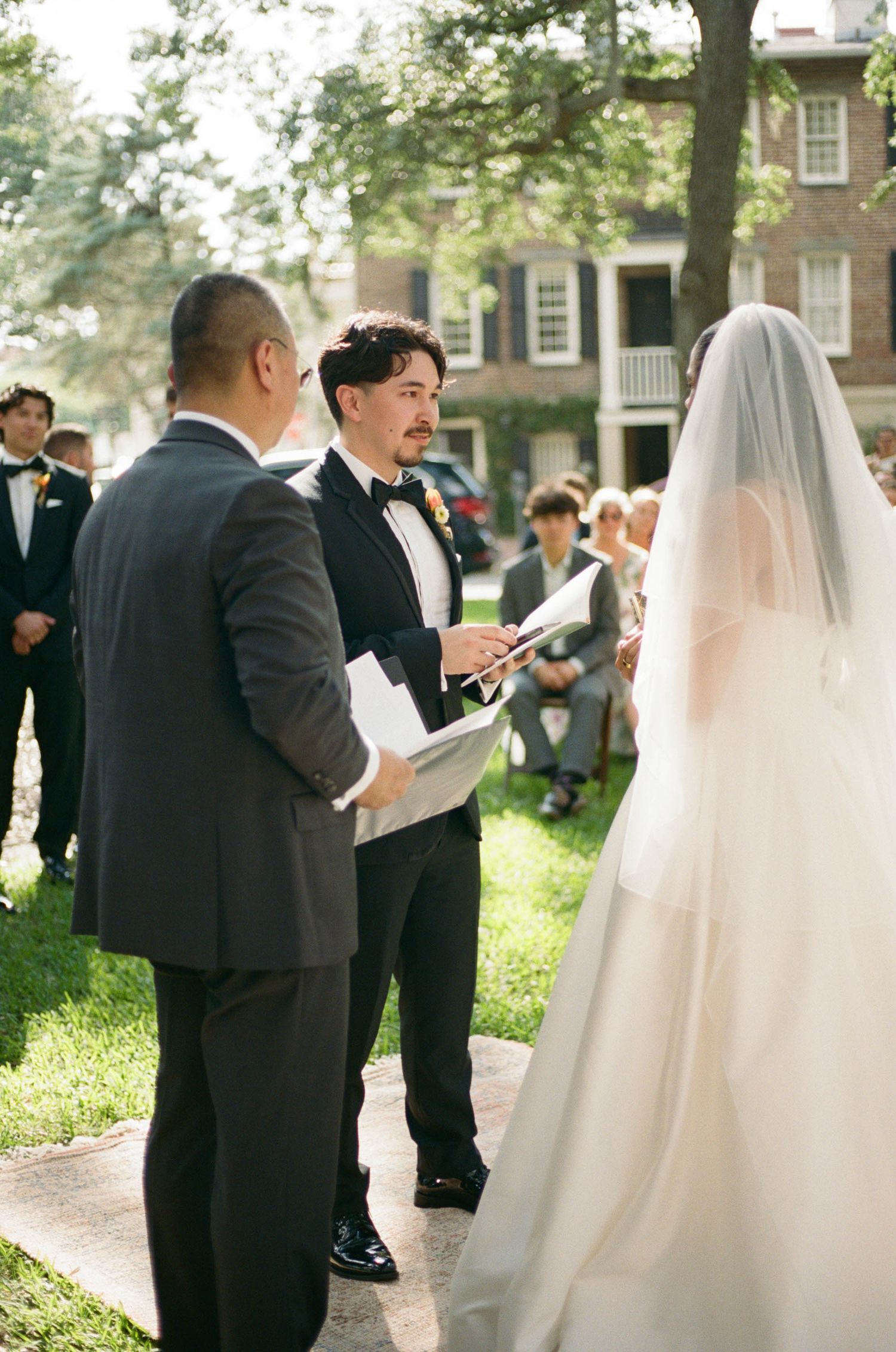 Wedding ceremony attendees gather outdoors under trees while reading from programs.